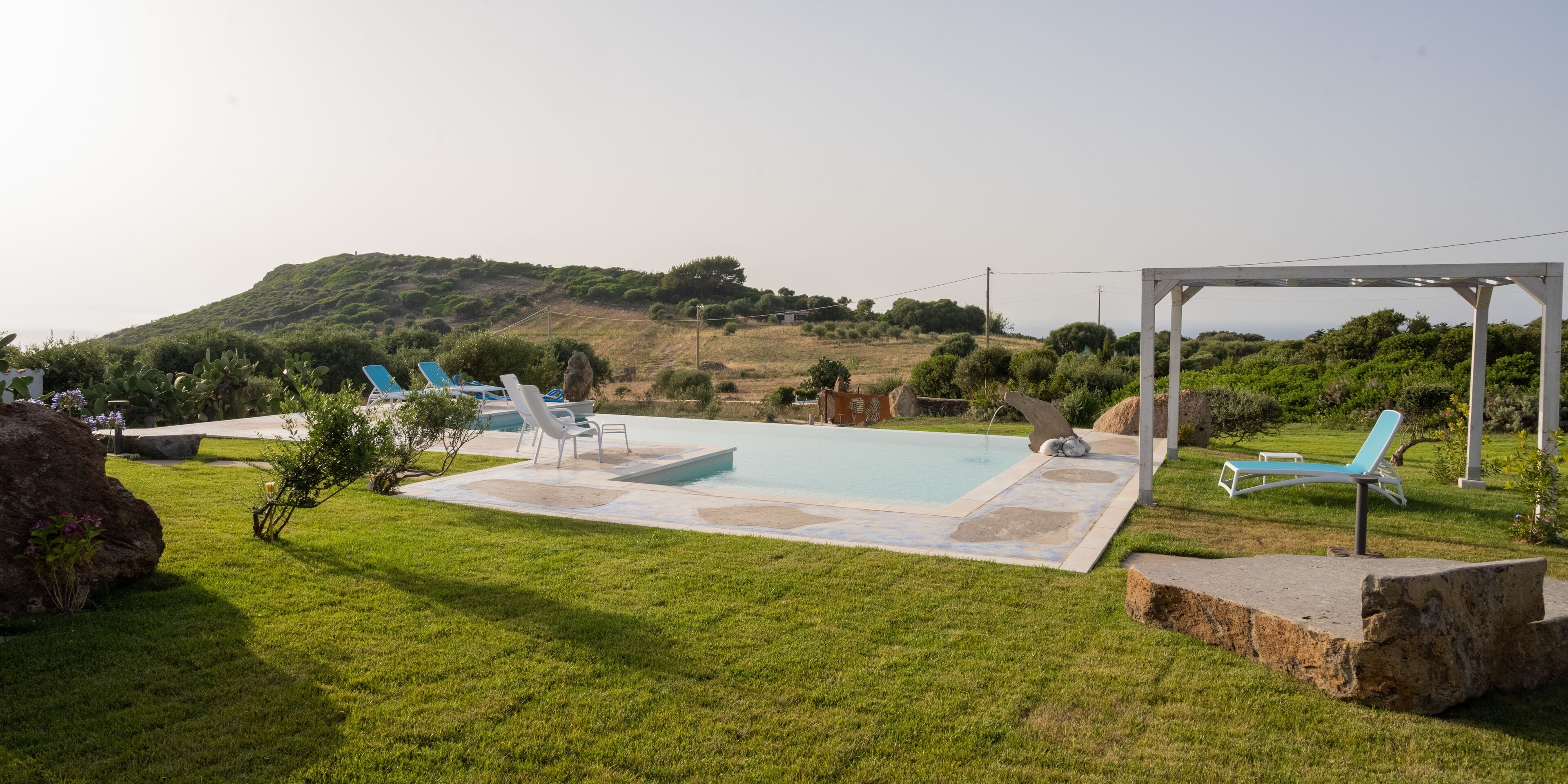 A pool area with lounge chairs, a pergola, and a view of a hilly landscape.