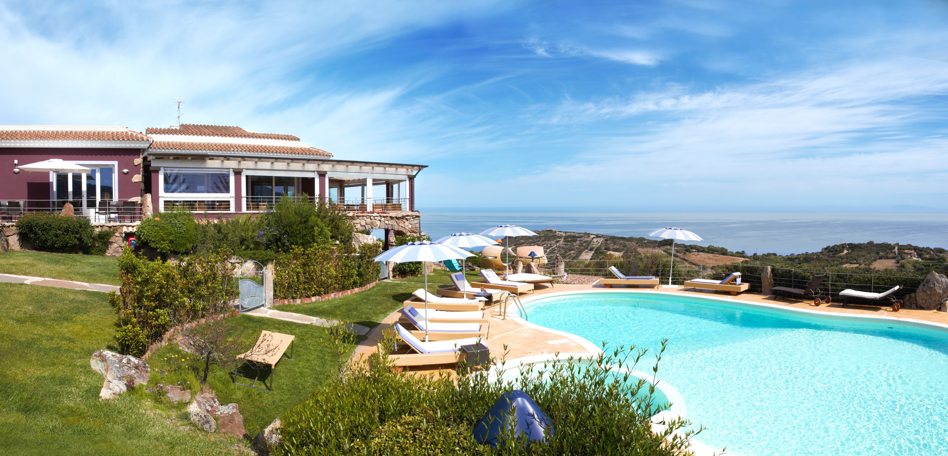 Una zona piscina con lettini e ombrelloni, una casa con terrazza e vista sul mare.