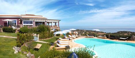 A pool area with lounge chairs and umbrellas, a house with a terrace, and a view of the sea.