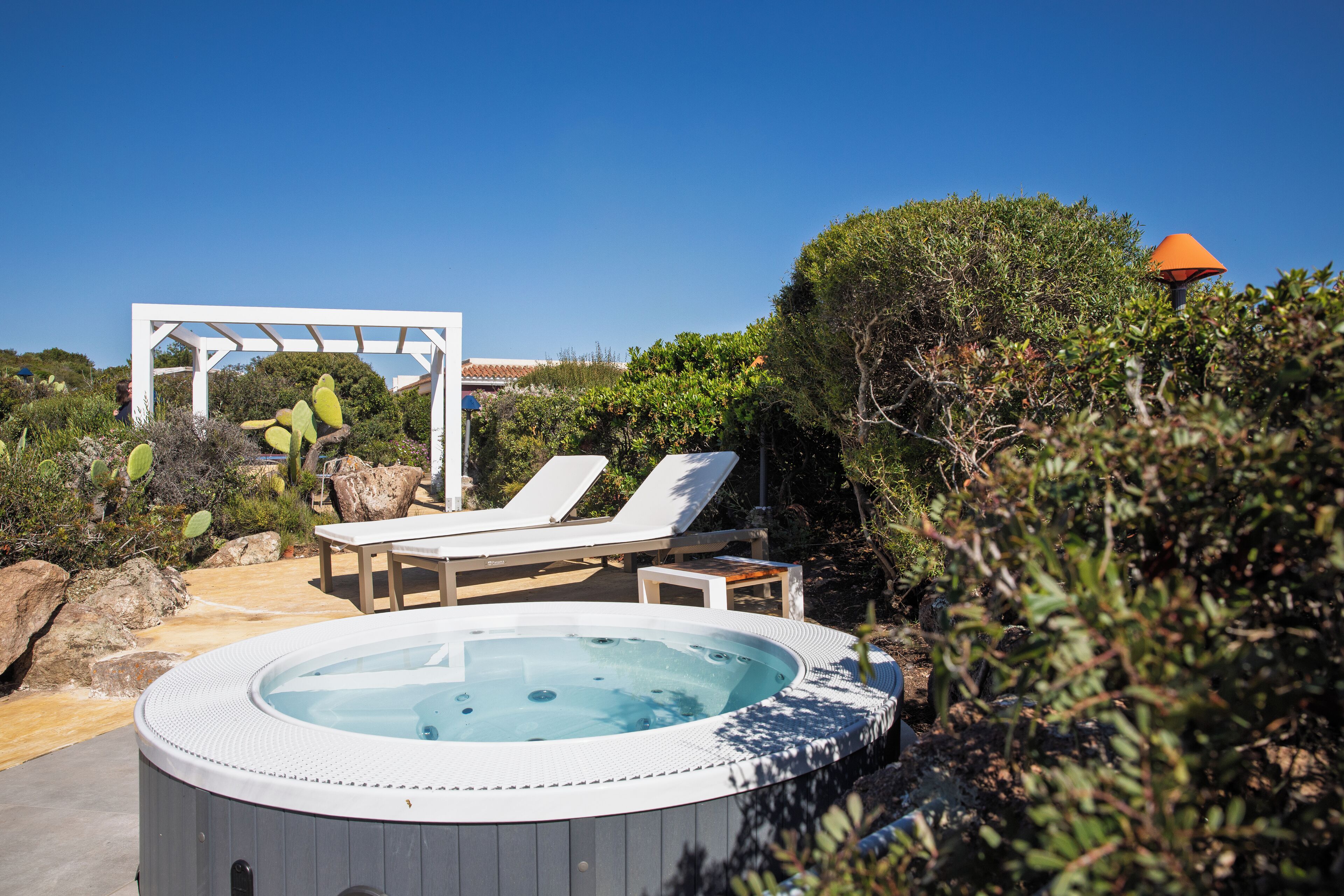 A hot tub surrounded by lounge chairs and a pergola, set in a garden with cacti and shrubs.