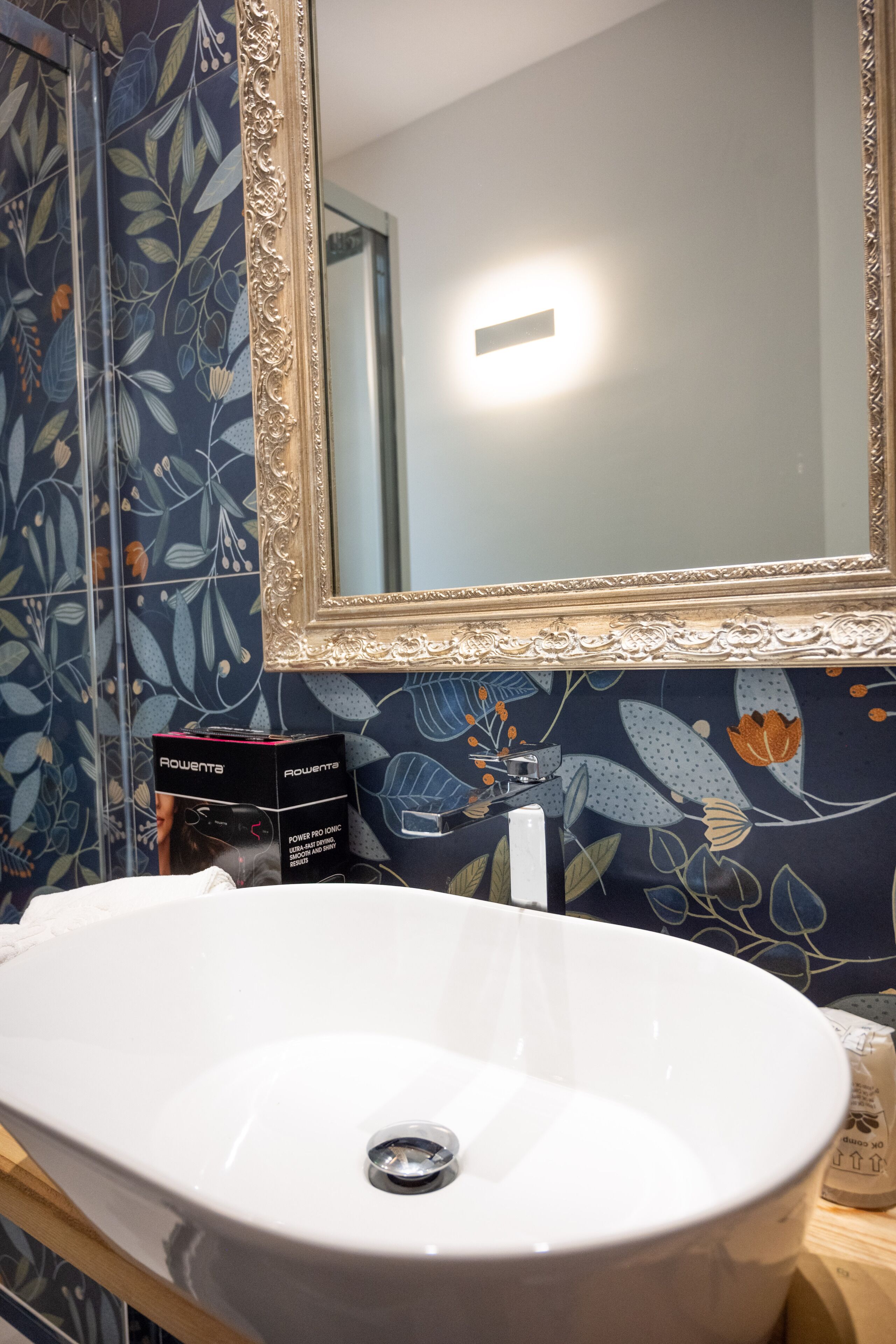 A bathroom with a white sink, a mirror with an ornate frame, and a floral-patterned wall.