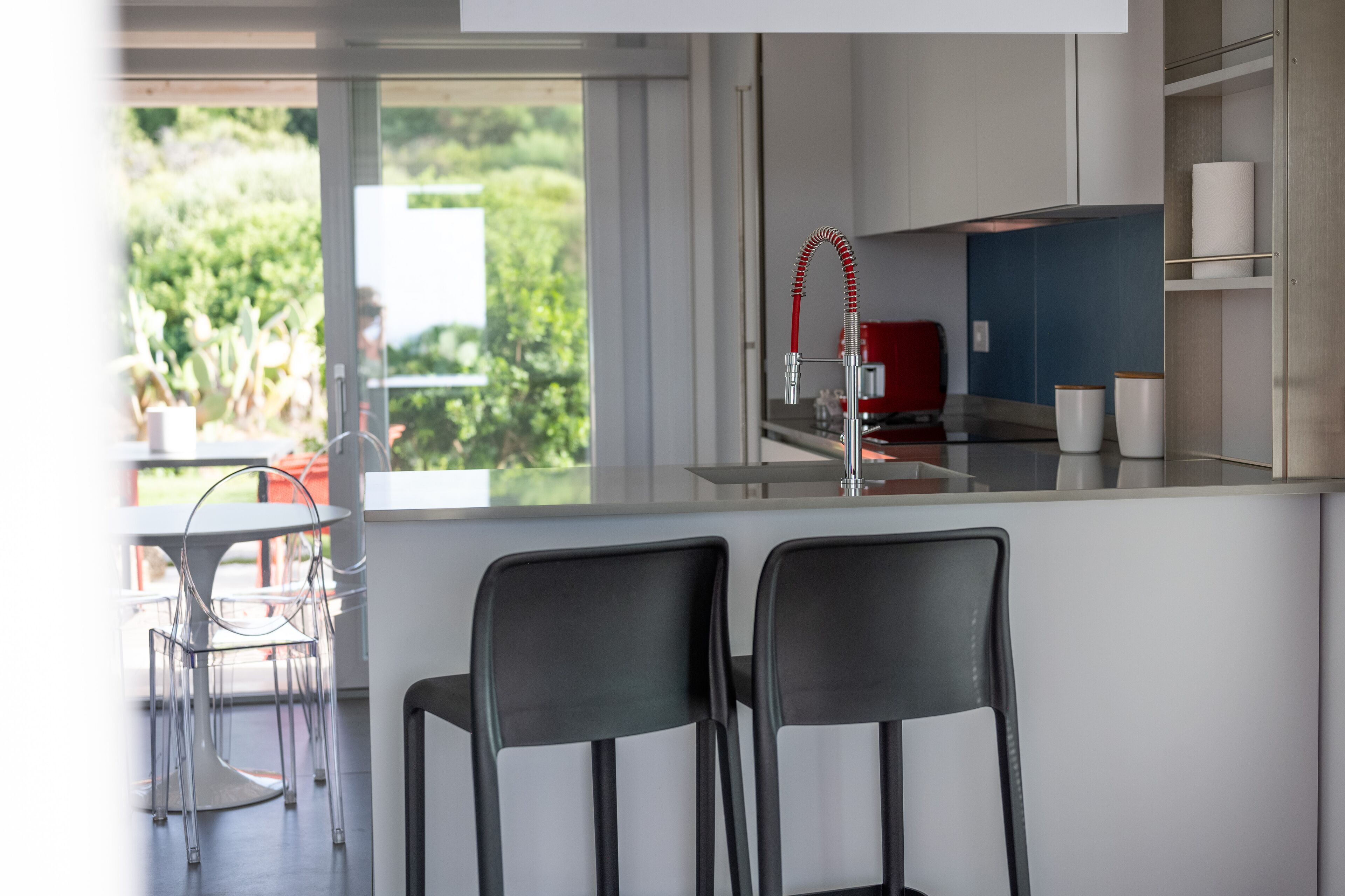 Una cucina moderna con una grande isola, due sedie nere, un rubinetto rosso e vista sul verde circostante.