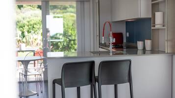 Une cuisine moderne dotée d’un grand îlot central, de deux chaises noires, d’un robinet rouge et offrant une vue sur la verdure à l’extérieur.