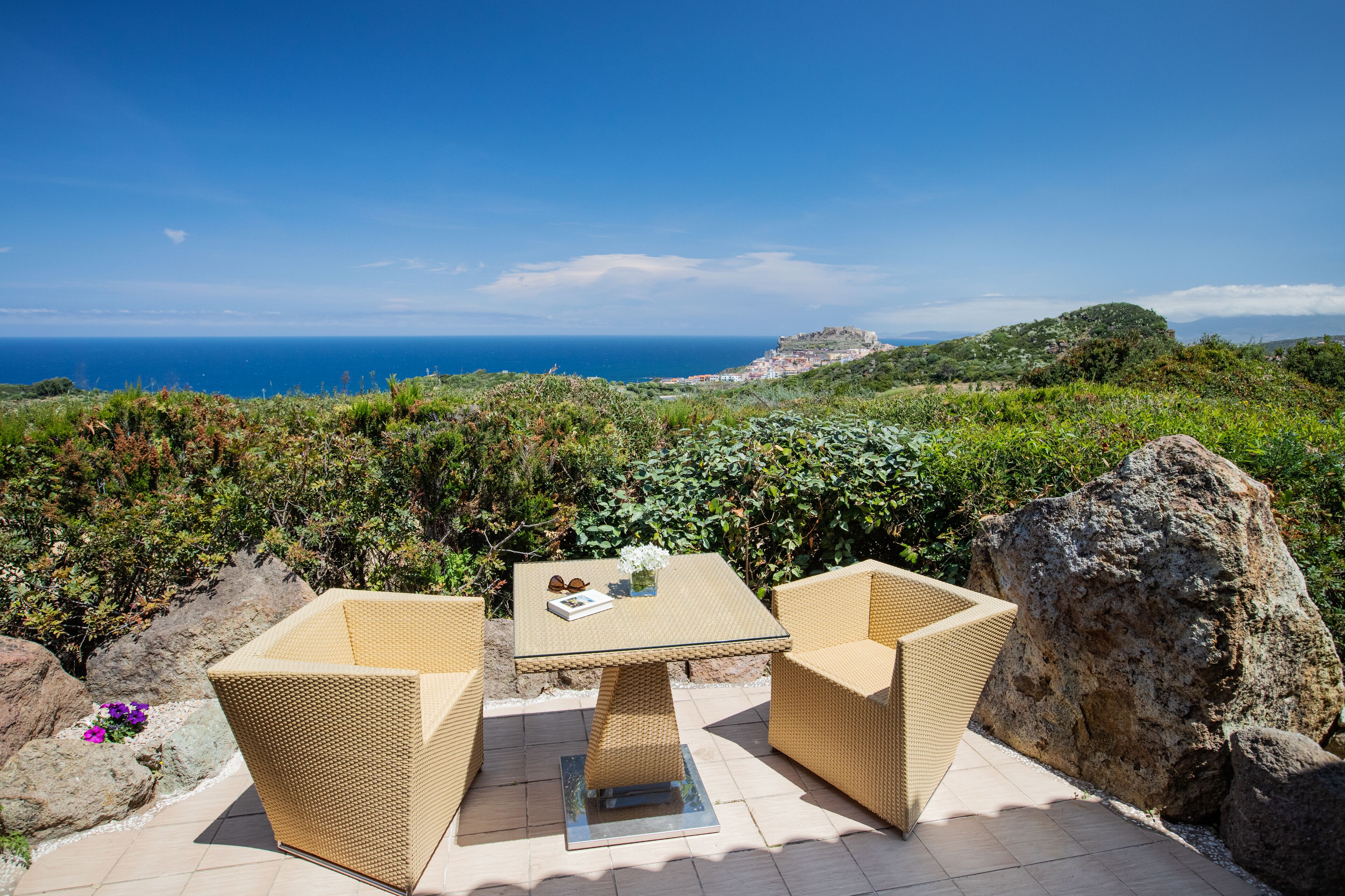 Un patio con mobiliario de mimbre y una mesa de vidrio, con vista a un paisaje costero y colinas lejanas junto al mar.
