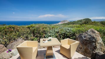 Un patio con mobiliario de mimbre y una mesa de vidrio, con vista a un paisaje costero y colinas lejanas junto al mar.