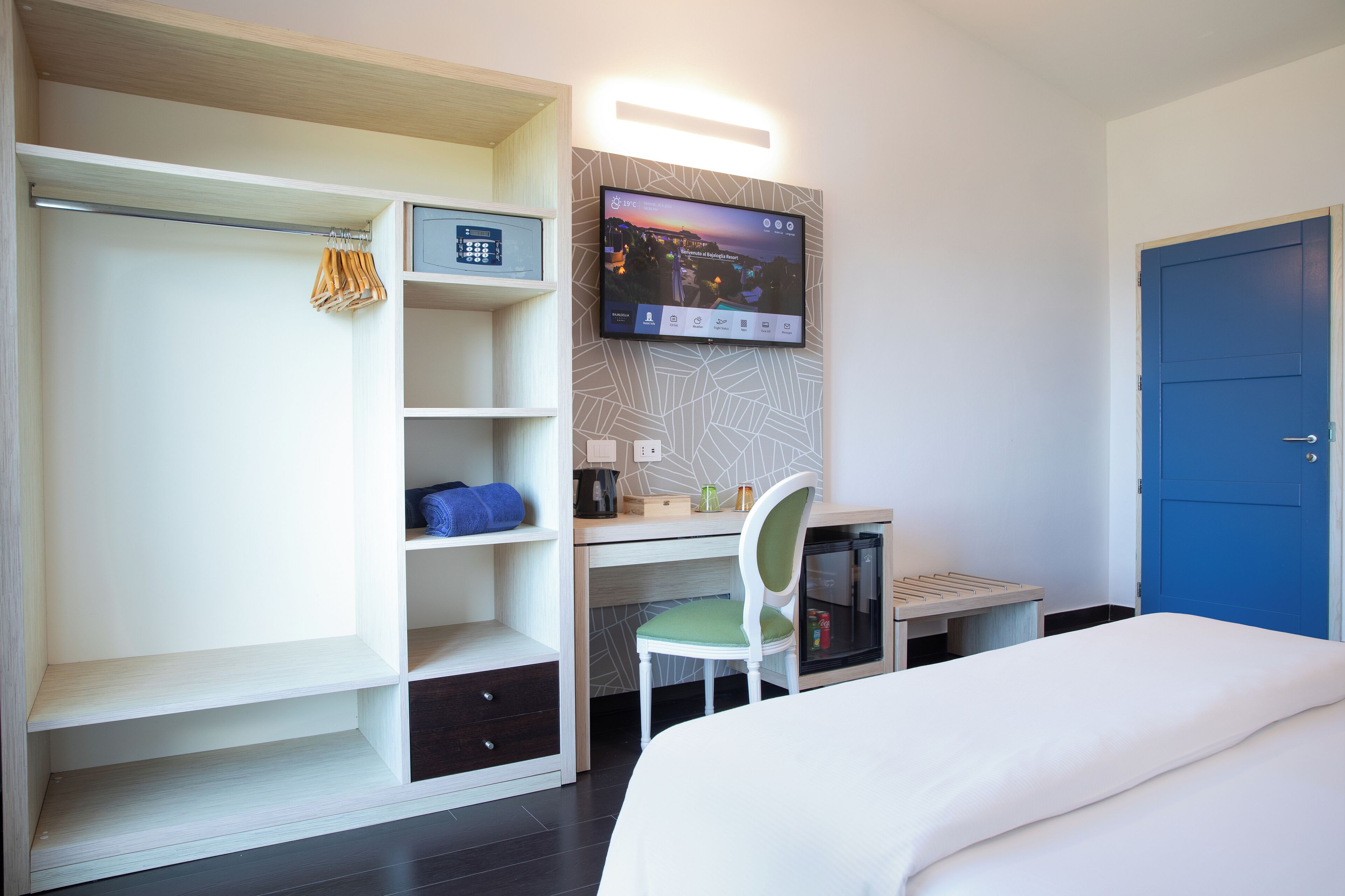 Une chambre d’hôtel avec un lit, un bureau et une chaise, un téléviseur fixé au mur, une armoire et une porte bleue.