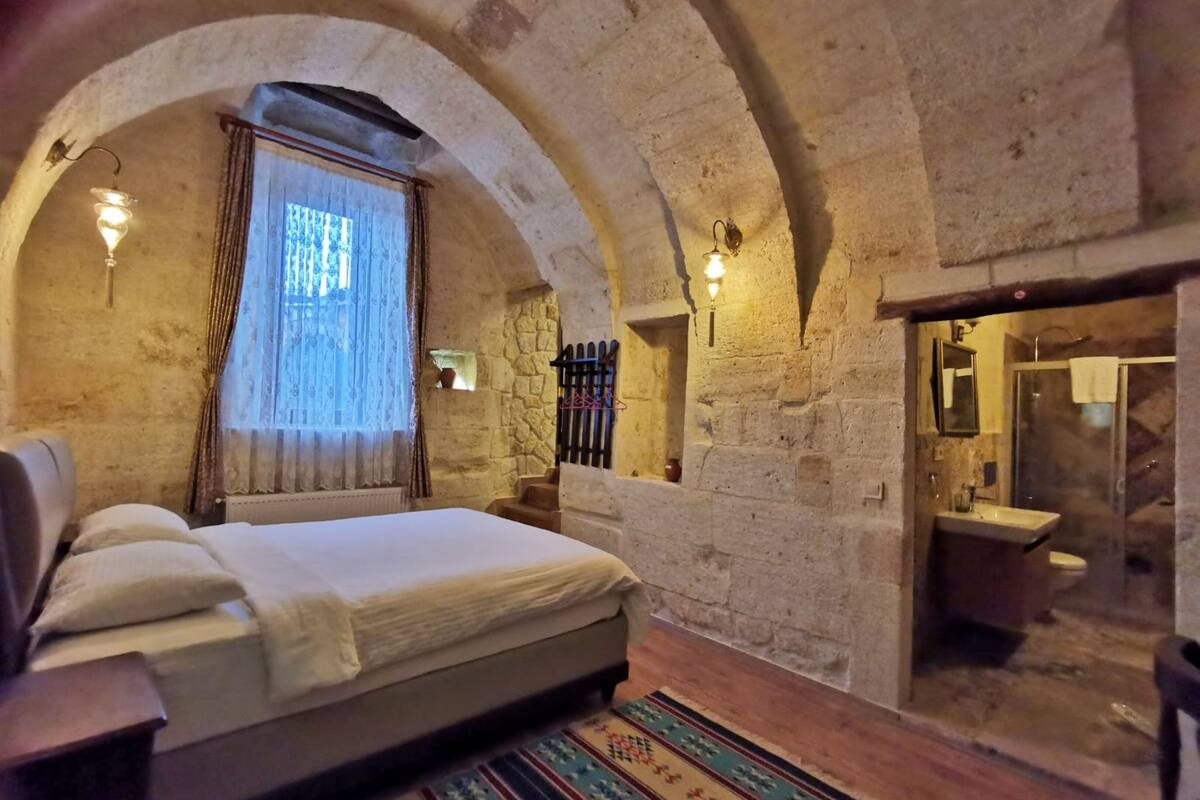 A room with a stone-walled arched ceiling, a bed with white linens, a wooden floor, and a bathroom with a sink and mirror.
