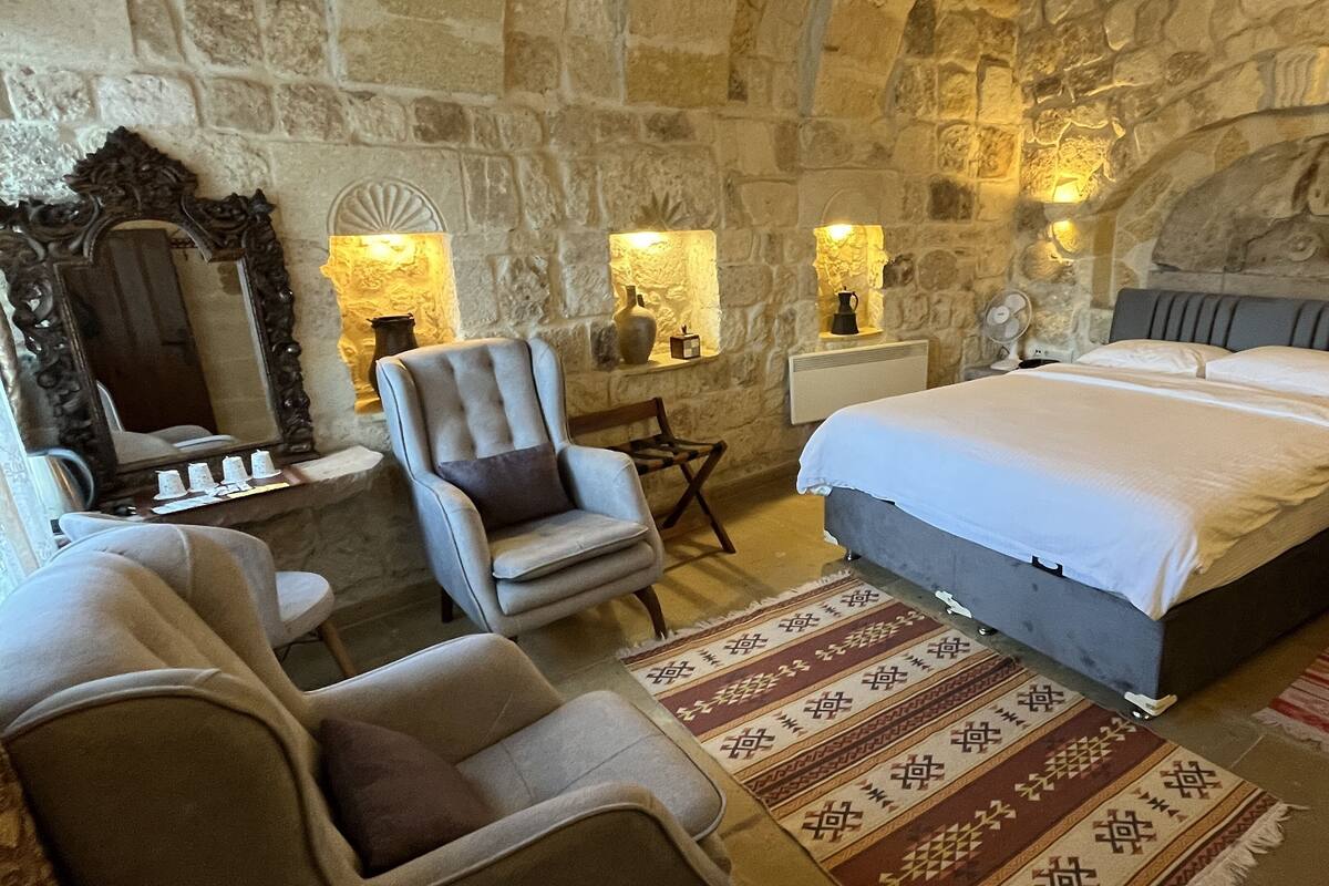 A bedroom with a stone wall, a bed with white linens, a chair, a small table, and a rug.