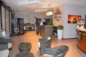 A modern coffee shop interior with wicker furniture, a counter with a menu, and a painting of a sunset on the wall. - Residence Europa (Alghero)