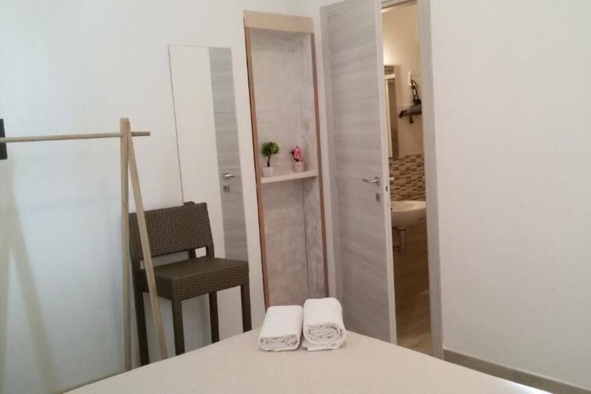 A hotel room with a bed, a chair, a small table with towels, a wardrobe, and a bathroom visible through an open door.