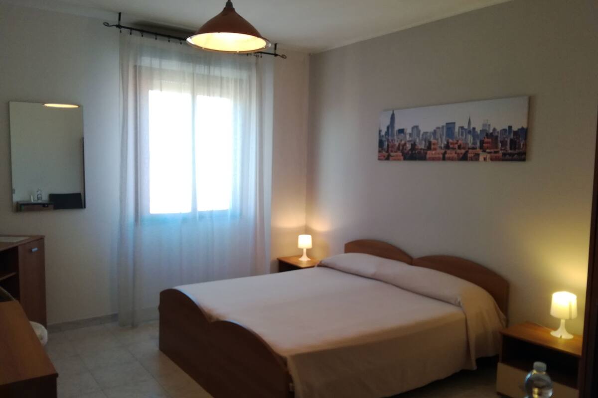 A hotel room with a bed, bedside tables, a mirror, a window with curtains, and a cityscape painting on the wall.