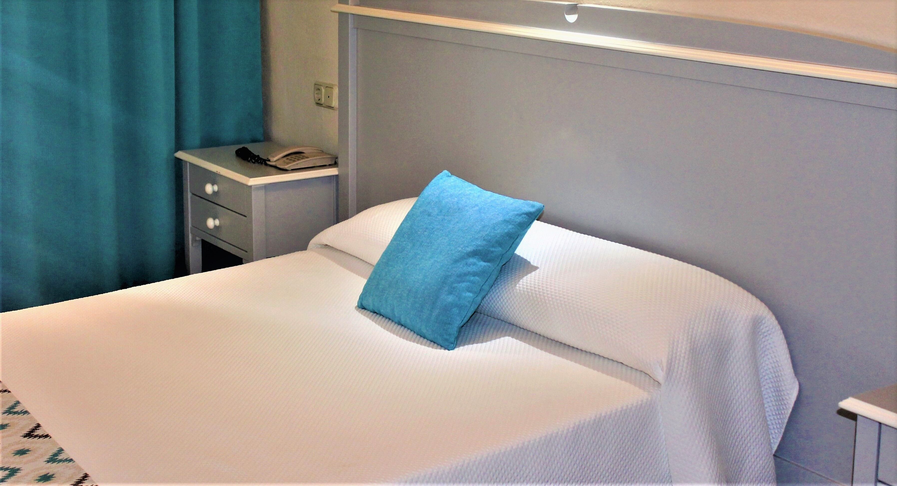 Una cama bien hecha con un único cojín azul, una mesita de noche con un teléfono y una cortina azul.