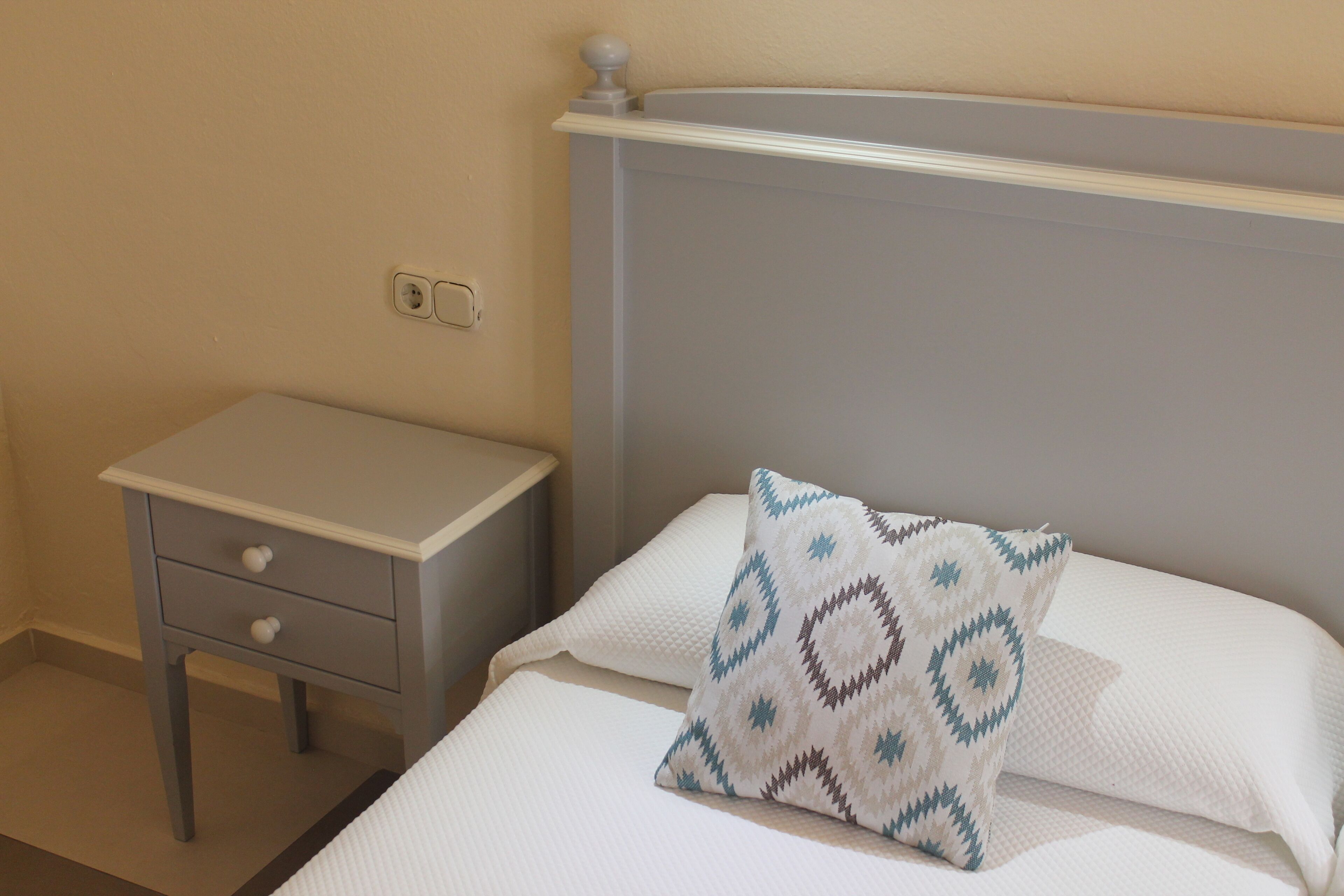 A neatly made bed with a patterned pillow and a small bedside table with drawers.