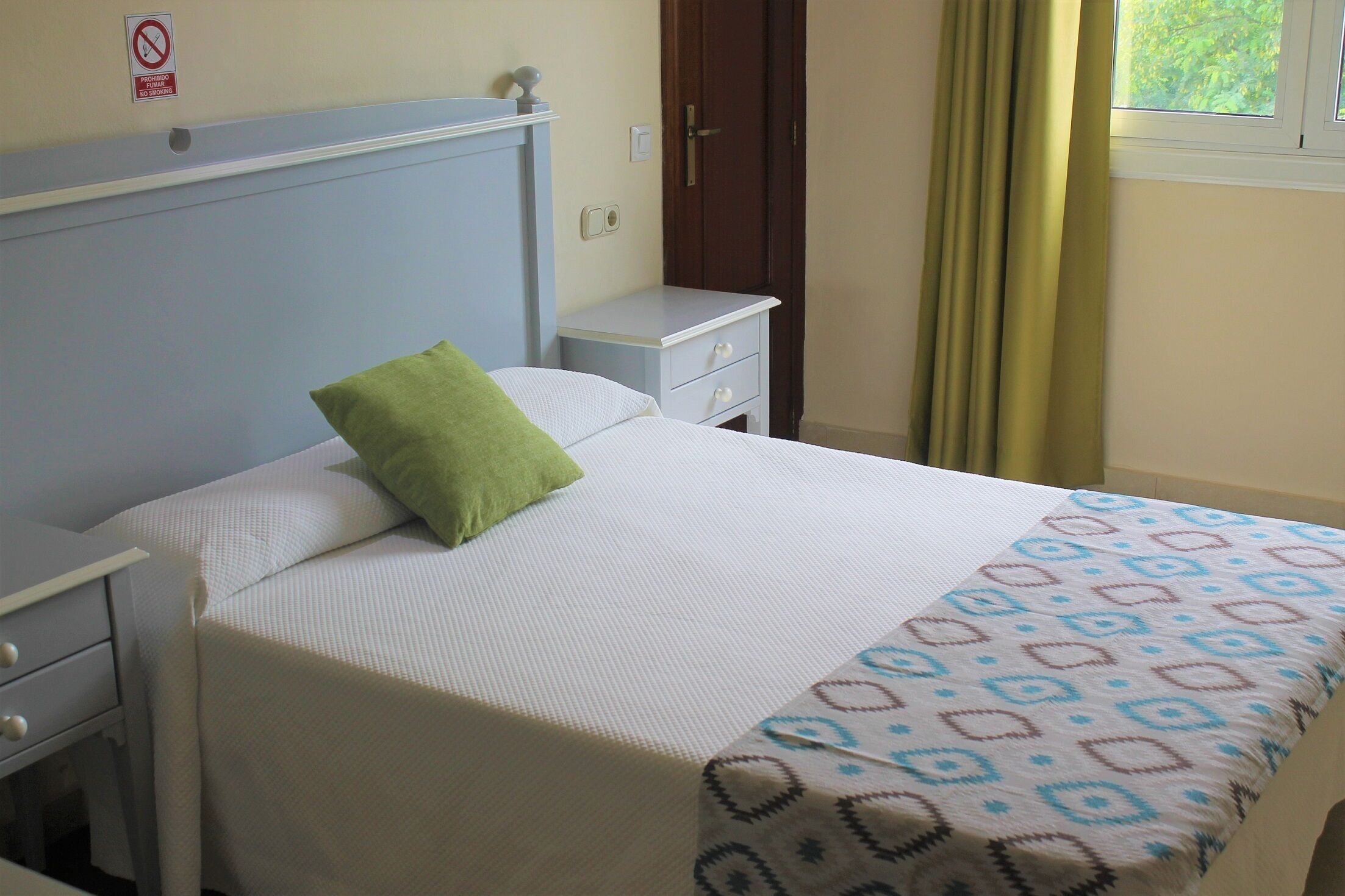 Una cama bien hecha con un cojín verde, una mesita de noche y una mesita auxiliar.