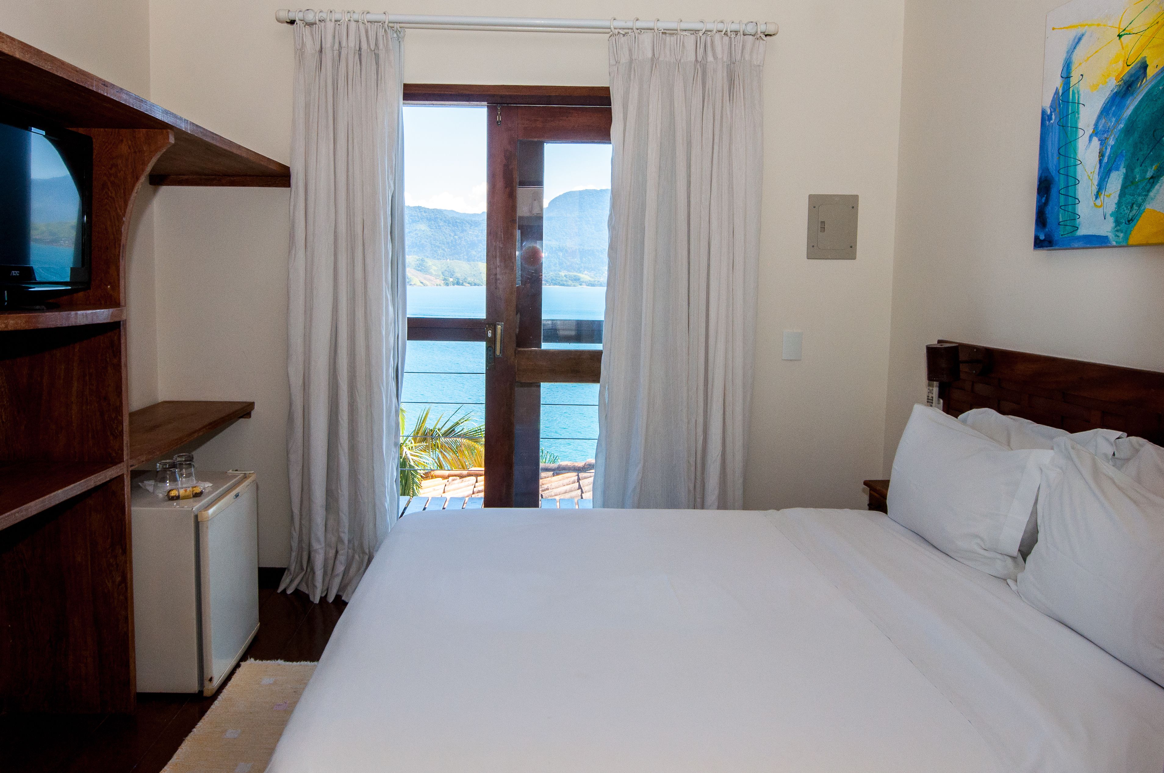 Un dormitorio con cama, televisión, refrigerador pequeño y vista al mar a través de una puerta.