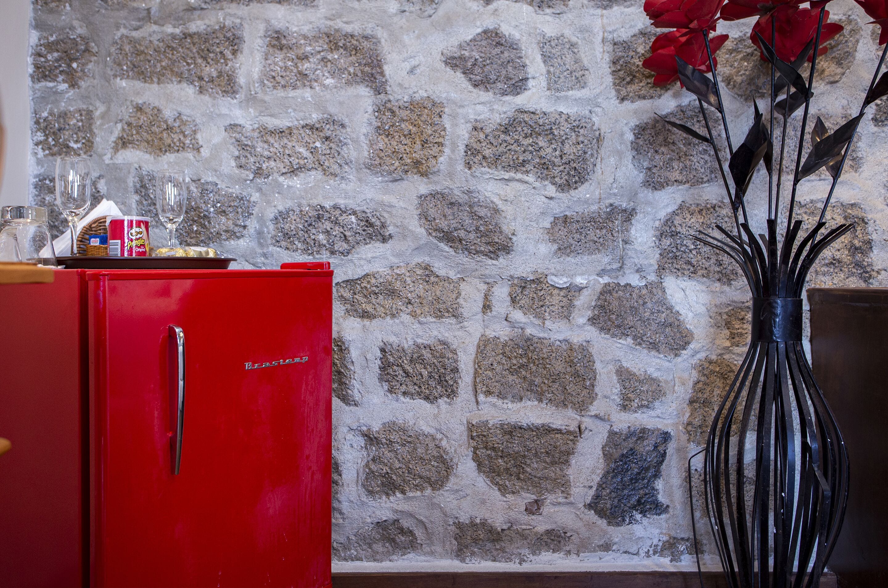 A red refrigerator with a tray of food and a bottle, a vase with red flowers, and a stone wall.
