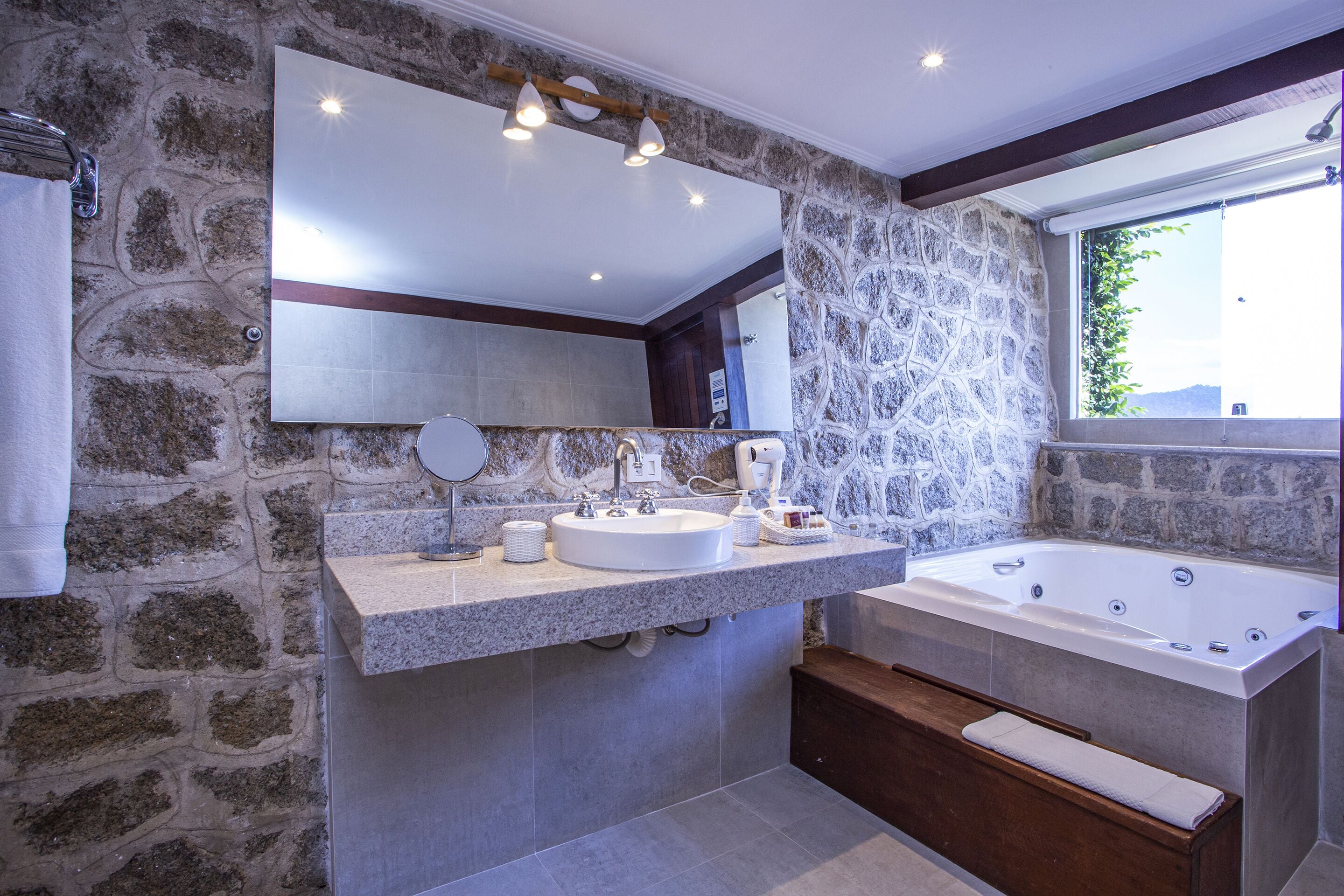 Un baño moderno con pared de piedra, un espejo grande, un lavabo, una bañera y una ventana que proporciona luz natural.