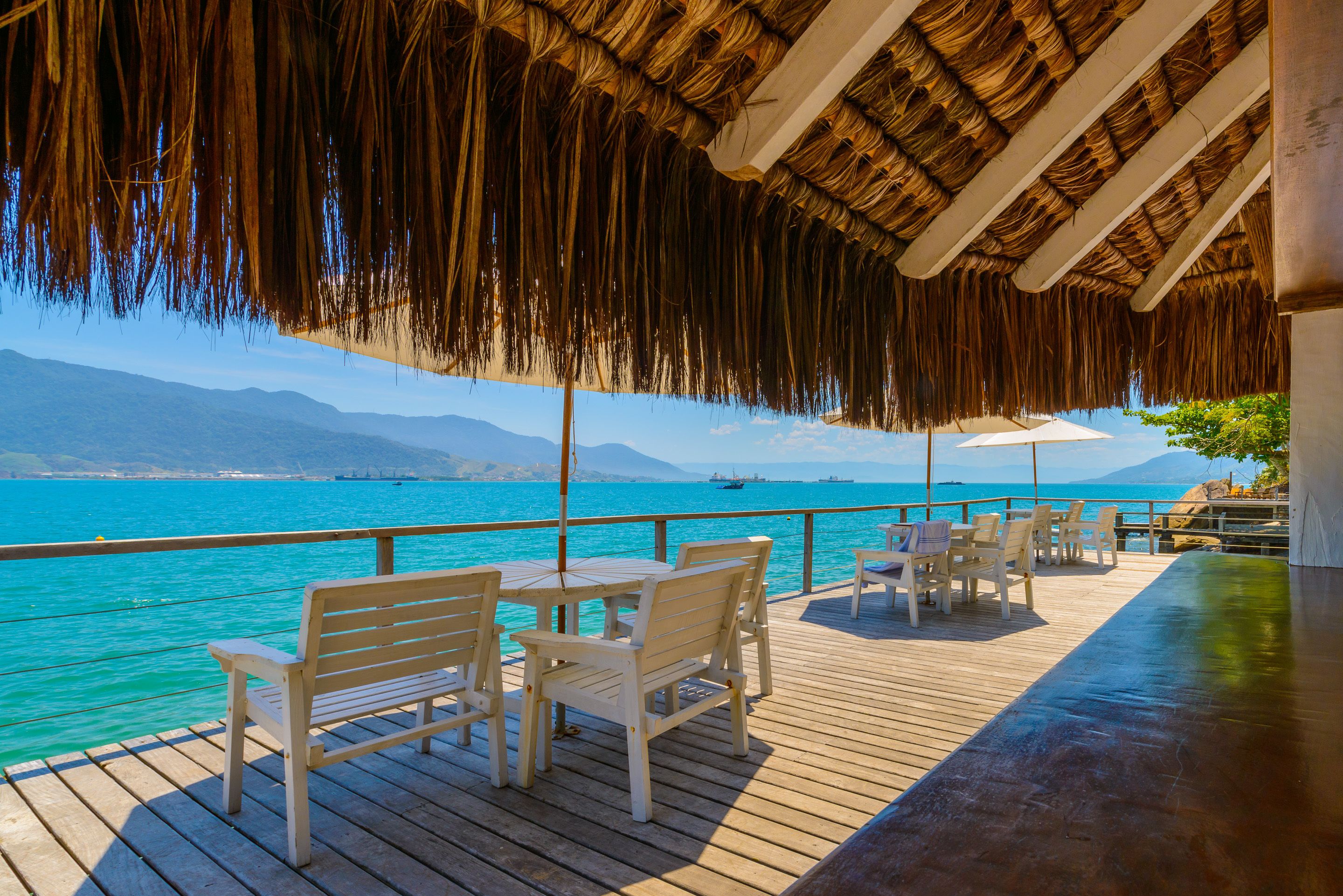 A wooden deck with white chairs and tables, overlooking a turquoise sea with a boat in the distance and mountains in the background.
