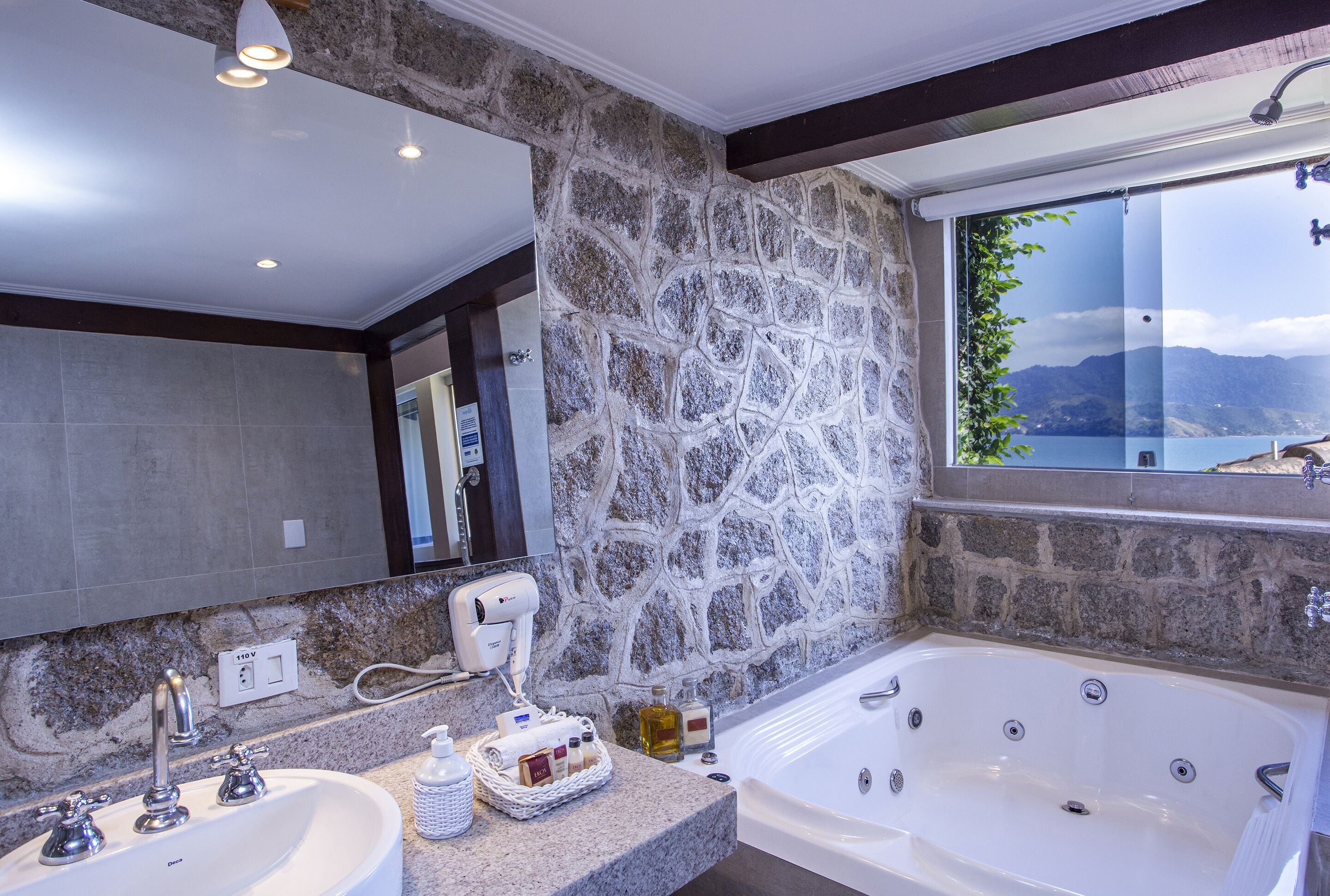 A bathroom with a stone wall, a large bathtub, and a view of mountains through a window.