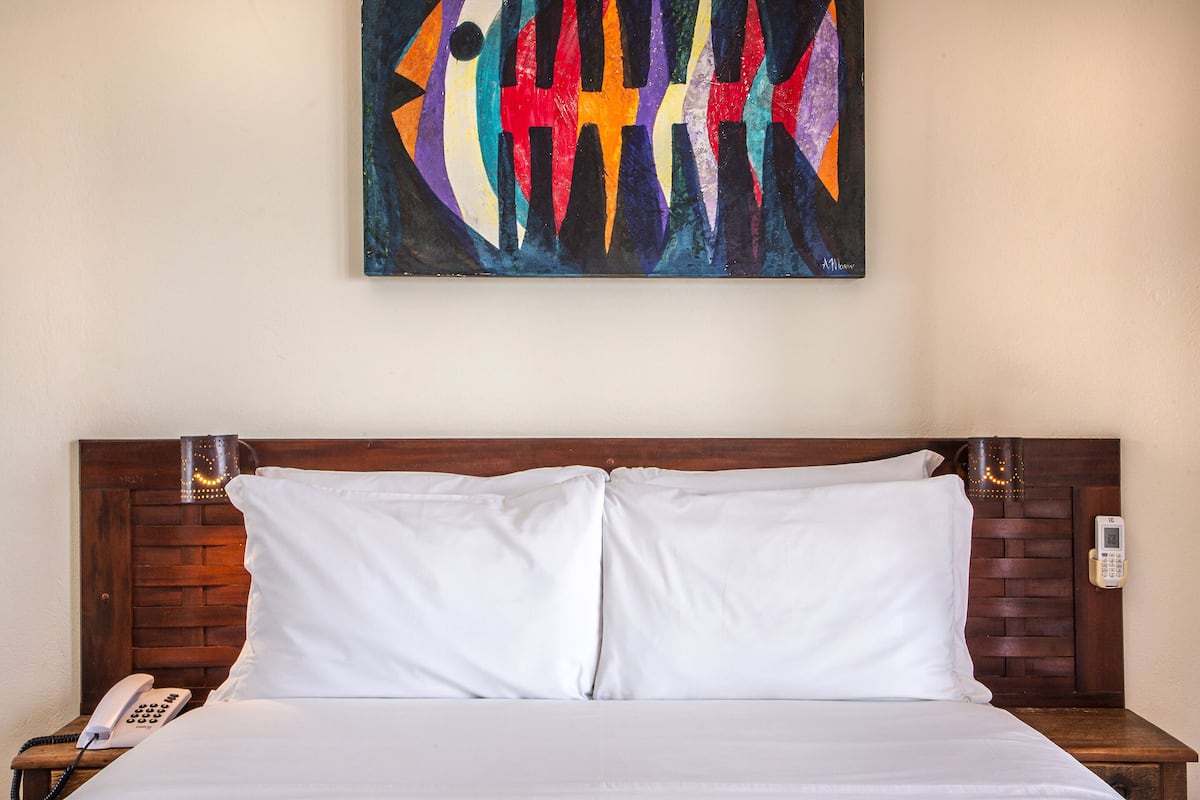 Een hotelkamer met een houten hoofdeinde, wit bedlinnen, een bedtelefoon en een kleurrijk abstract schilderij aan de muur.