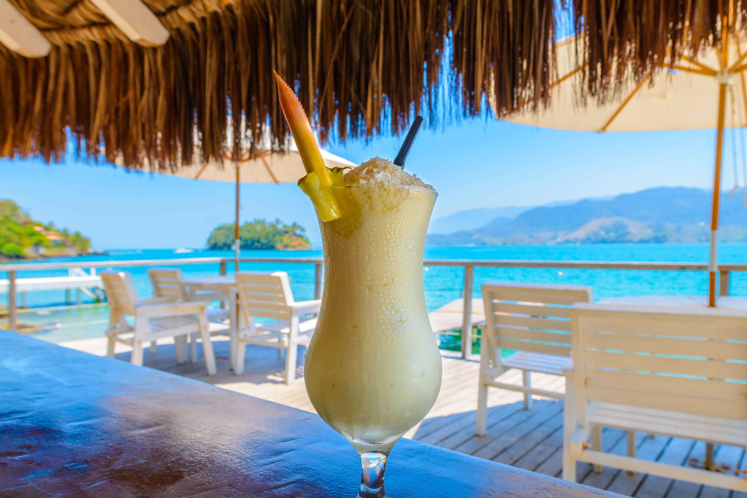 A drink with a straw and a fruit garnish on a table by a poolside with chairs and a thatched roof.