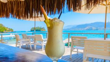 A drink with a straw and a fruit garnish on a table by a poolside with chairs and a thatched roof.