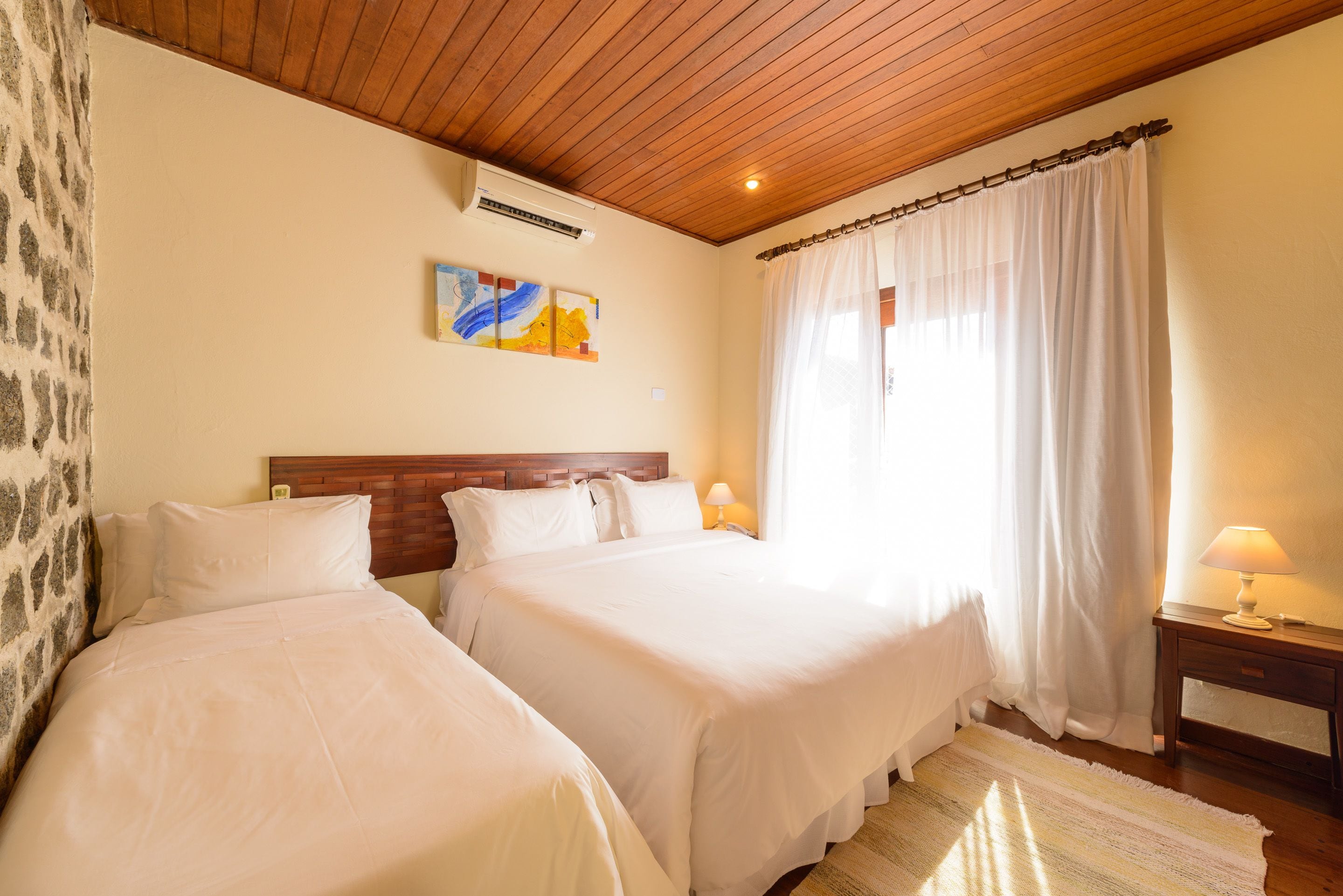 A hotel room with two beds, wooden ceiling, white curtains, a painting on the wall, and a bedside table with a lamp.
