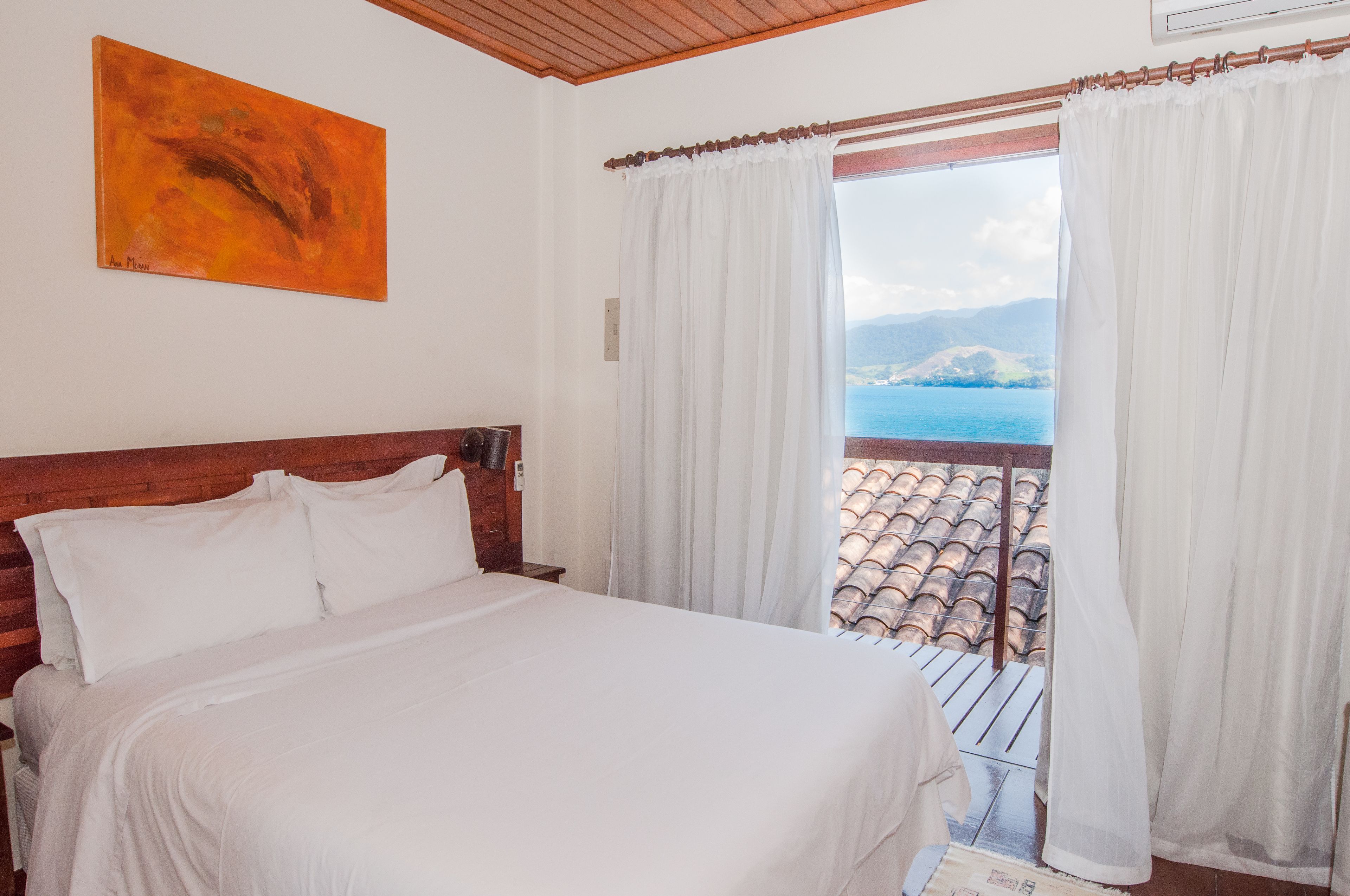 Un dormitorio con cabecera de madera, una cama grande, ropa de cama blanca, un cuadro en la pared y un balcón con cortinas blancas y vista al mar.