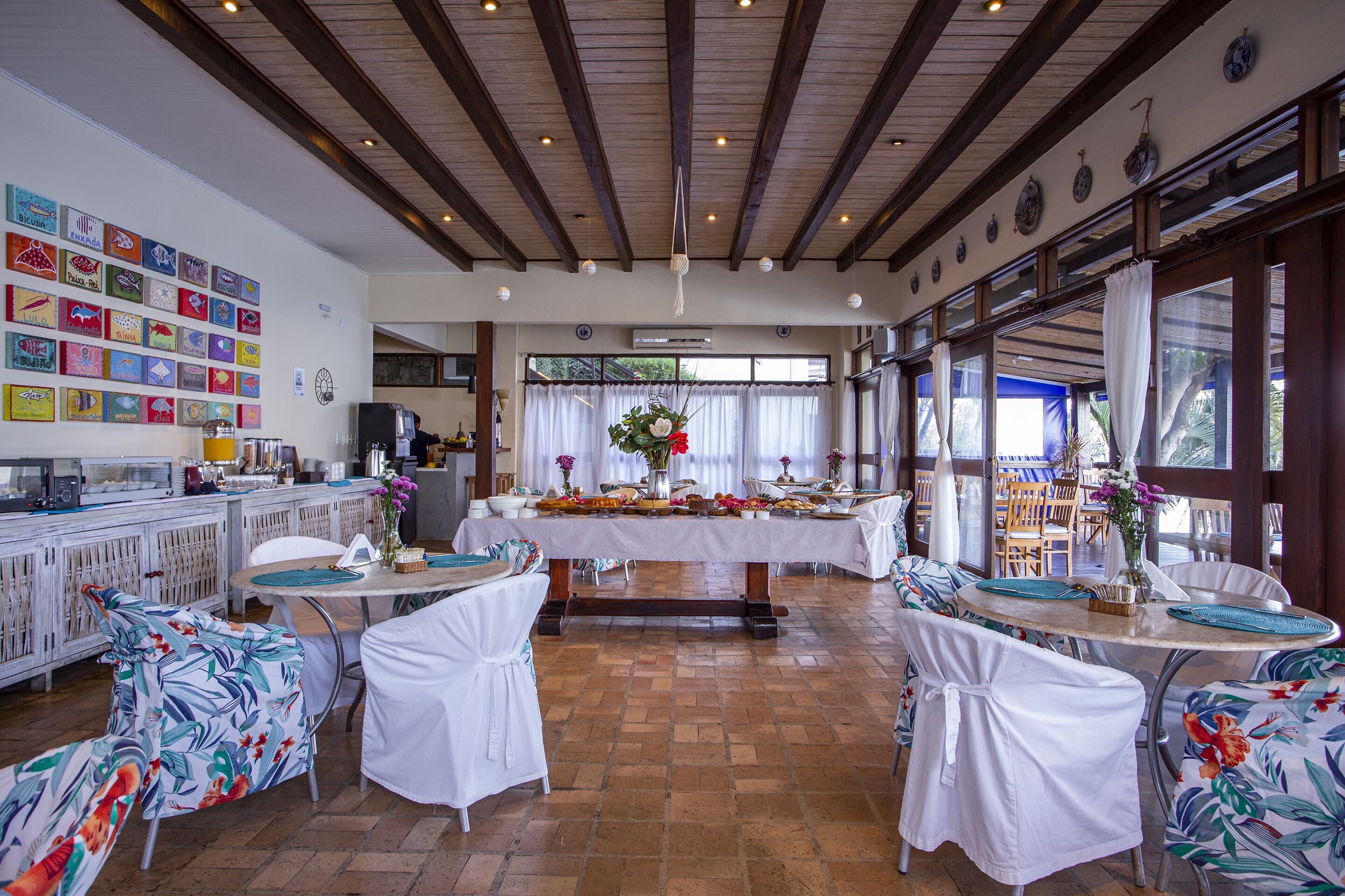 Un restaurant avec une longue table à manger, des chaises blanches et un espace cuisine à l’arrière-plan.