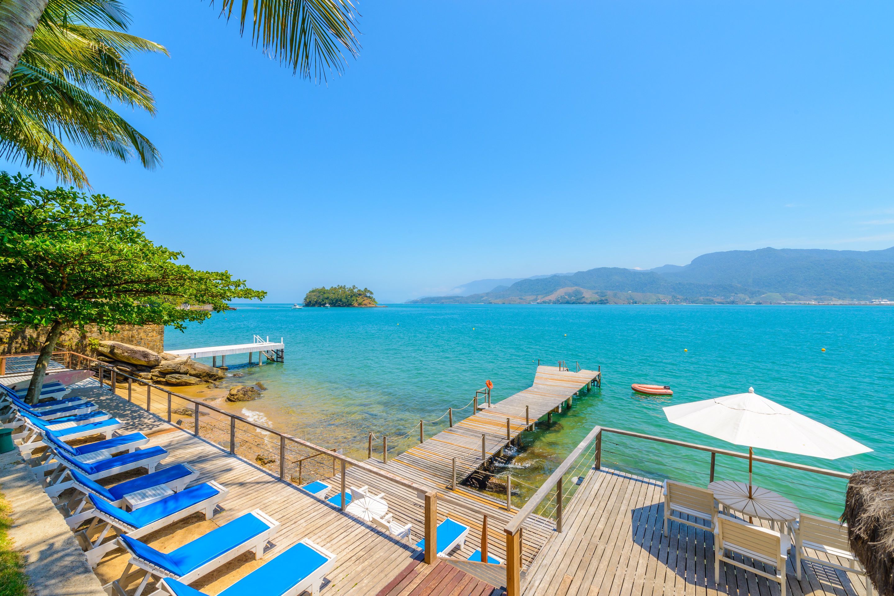 A wooden deck with blue lounge chairs and a white umbrella, overlooking a clear blue sea with a small island and mountains in the distance.