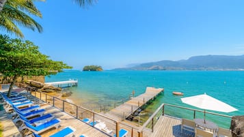 A wooden deck with blue lounge chairs and a white umbrella, overlooking a clear blue sea with a small island and mountains in the distance.