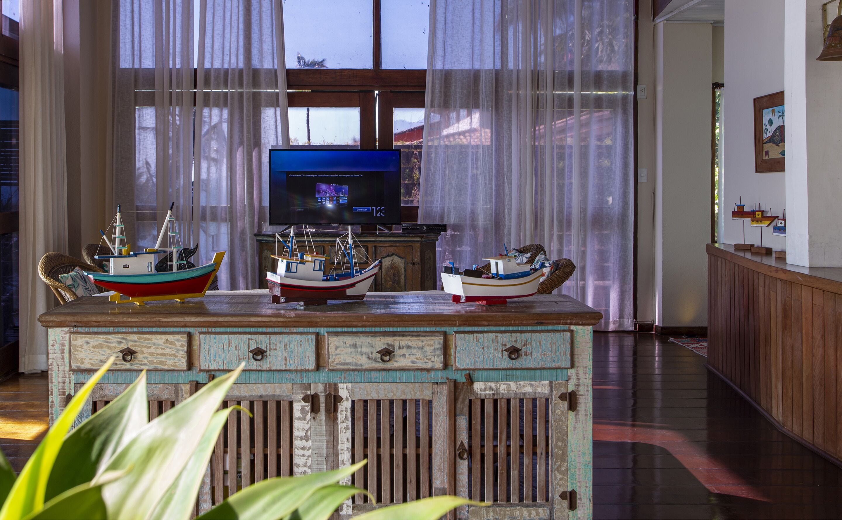 A room with a wooden cabinet displaying model boats, a television on a stand, and a window with sheer curtains.