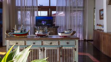 Una habitación con un aparador de madera que exhibe barcos modelo, un televisor sobre un soporte y una ventana con cortinas traslúcidas.