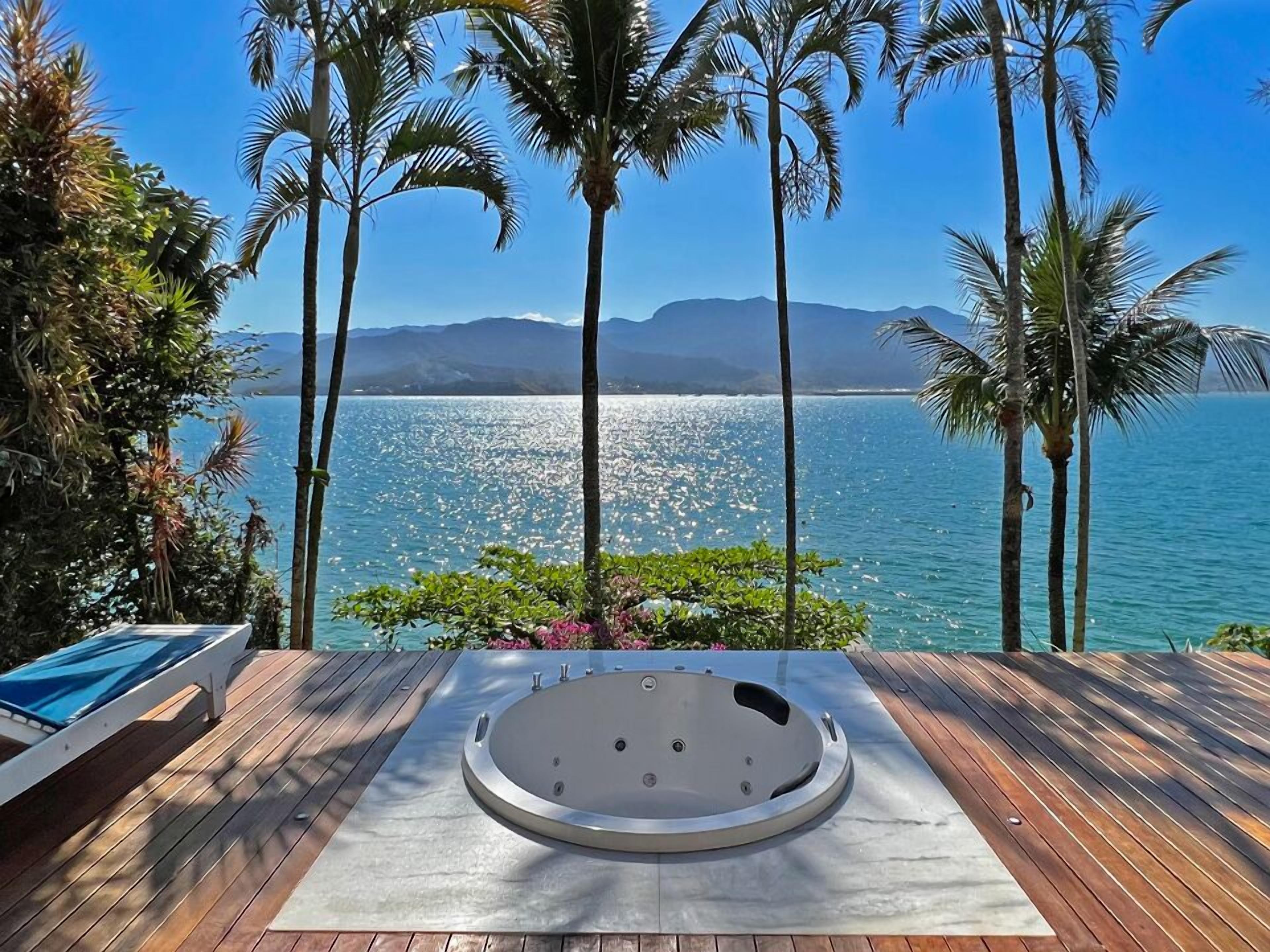 A wooden deck with a hot tub, surrounded by palm trees and overlooking a body of water with mountains in the background.