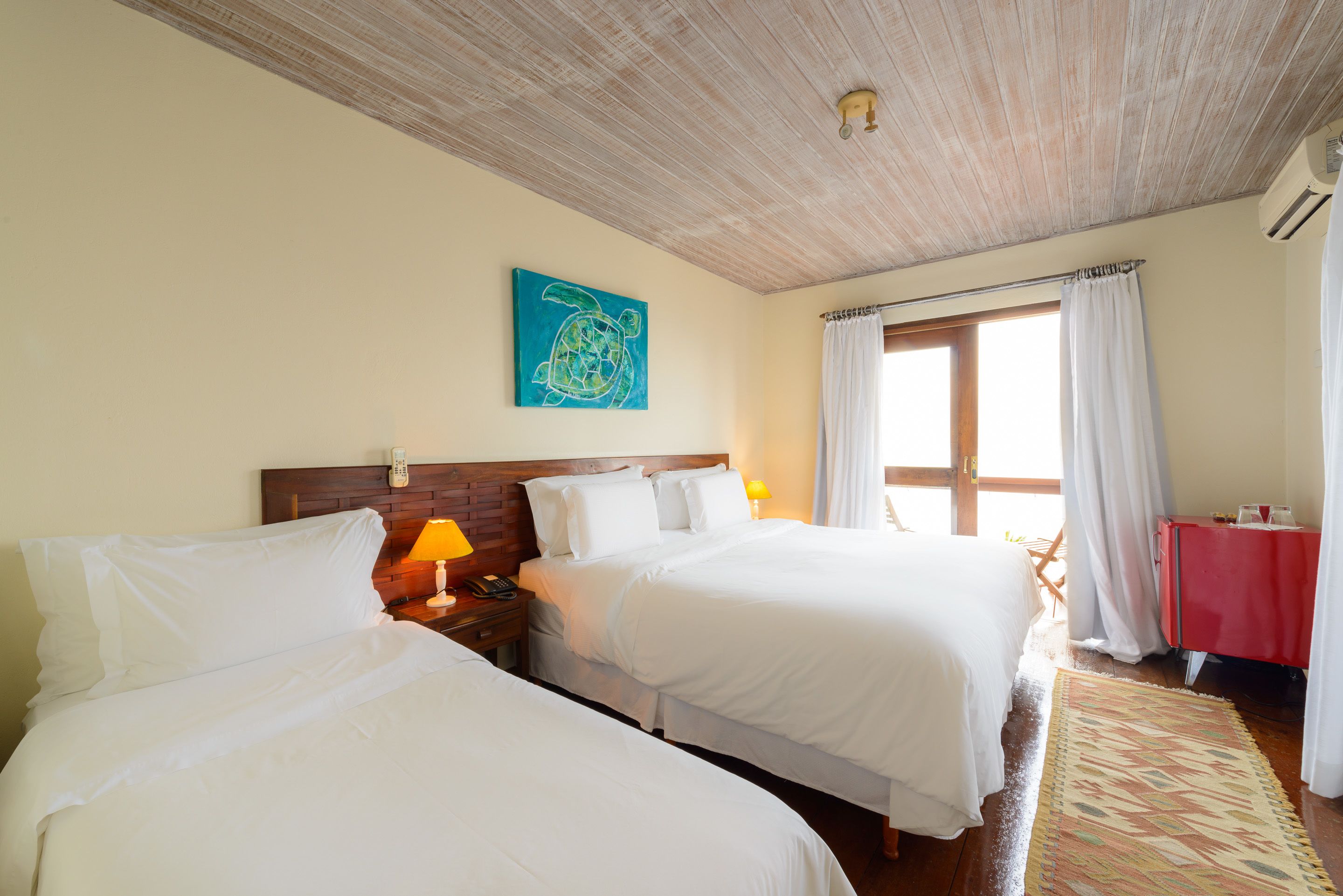 A hotel room with two beds, a wooden ceiling, a blue artwork, a window with curtains, and a red table.