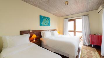 A hotel room with two beds, a wooden ceiling, a blue artwork, a window with curtains, and a red table.