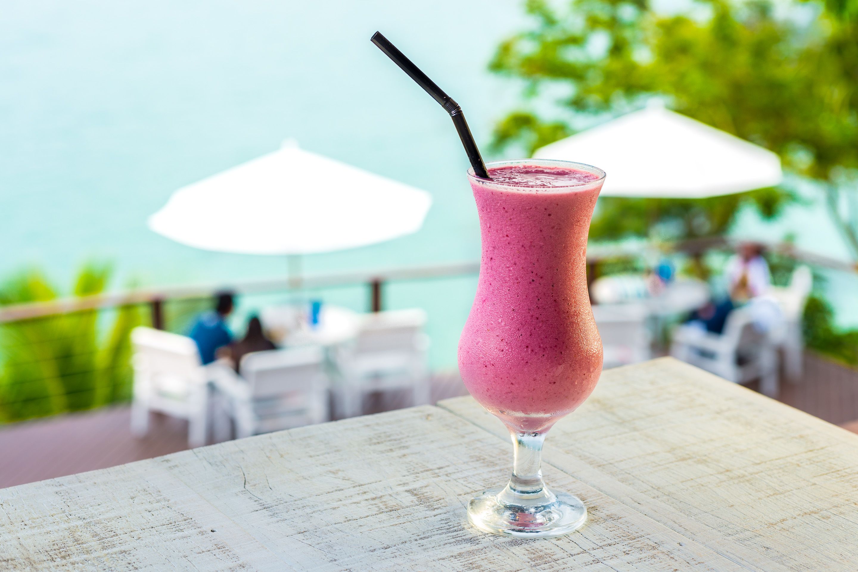 A pink smoothie in a tall glass with a straw, placed on a table with a view of a beach and umbrellas in the background.