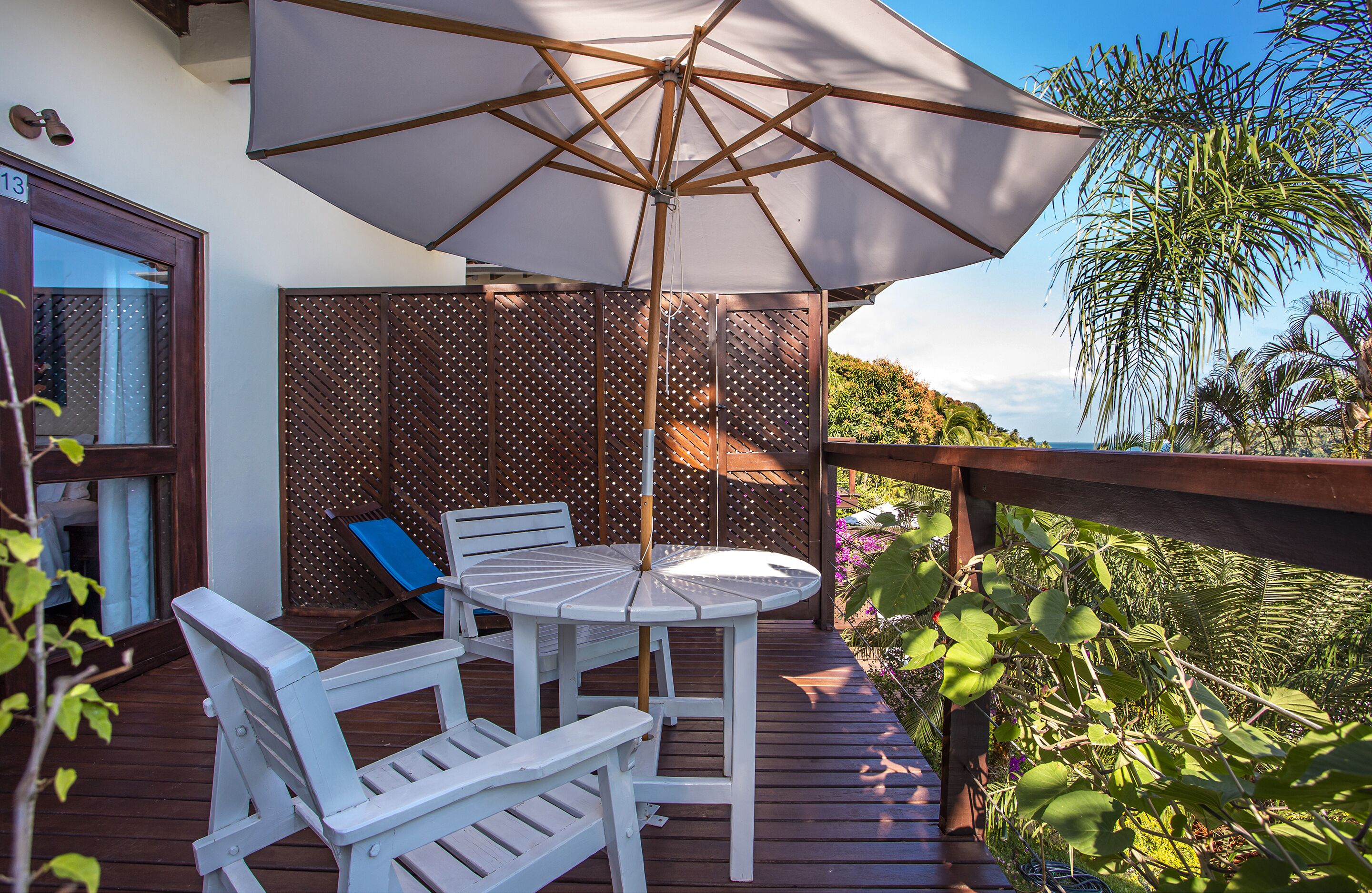 A wooden deck with a table and chairs under a large umbrella, surrounded by greenery and a view of the ocean.