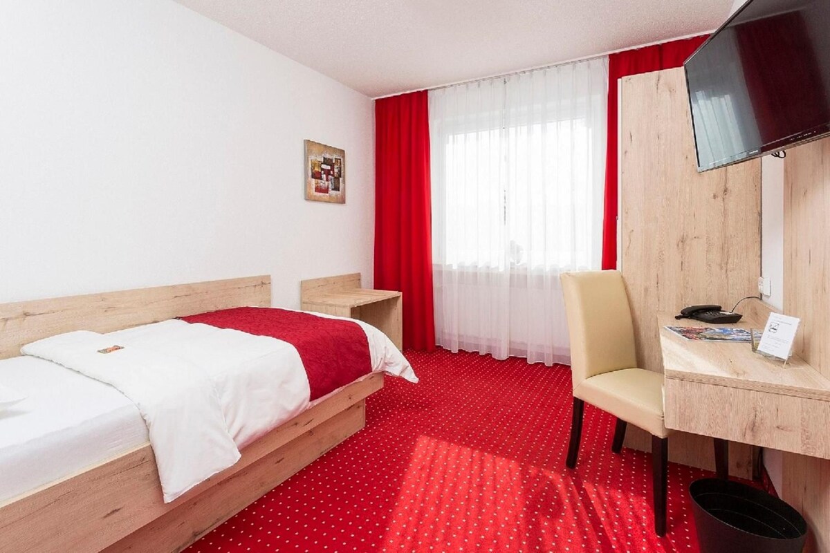 A hotel room with a bed, a desk, a chair, a TV, and a window with curtains.