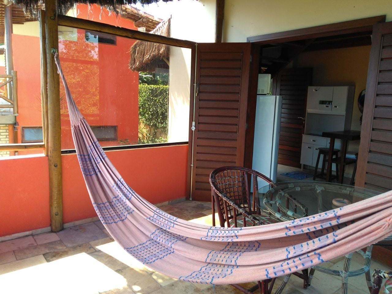 A hammock in a sunlit outdoor area with a glass table and chairs, a wooden door, and a view of a building with a red exterior.