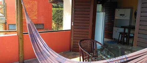 A hammock in a sunlit outdoor area with a glass table and chairs, a wooden door, and a view of a building with a red exterior.