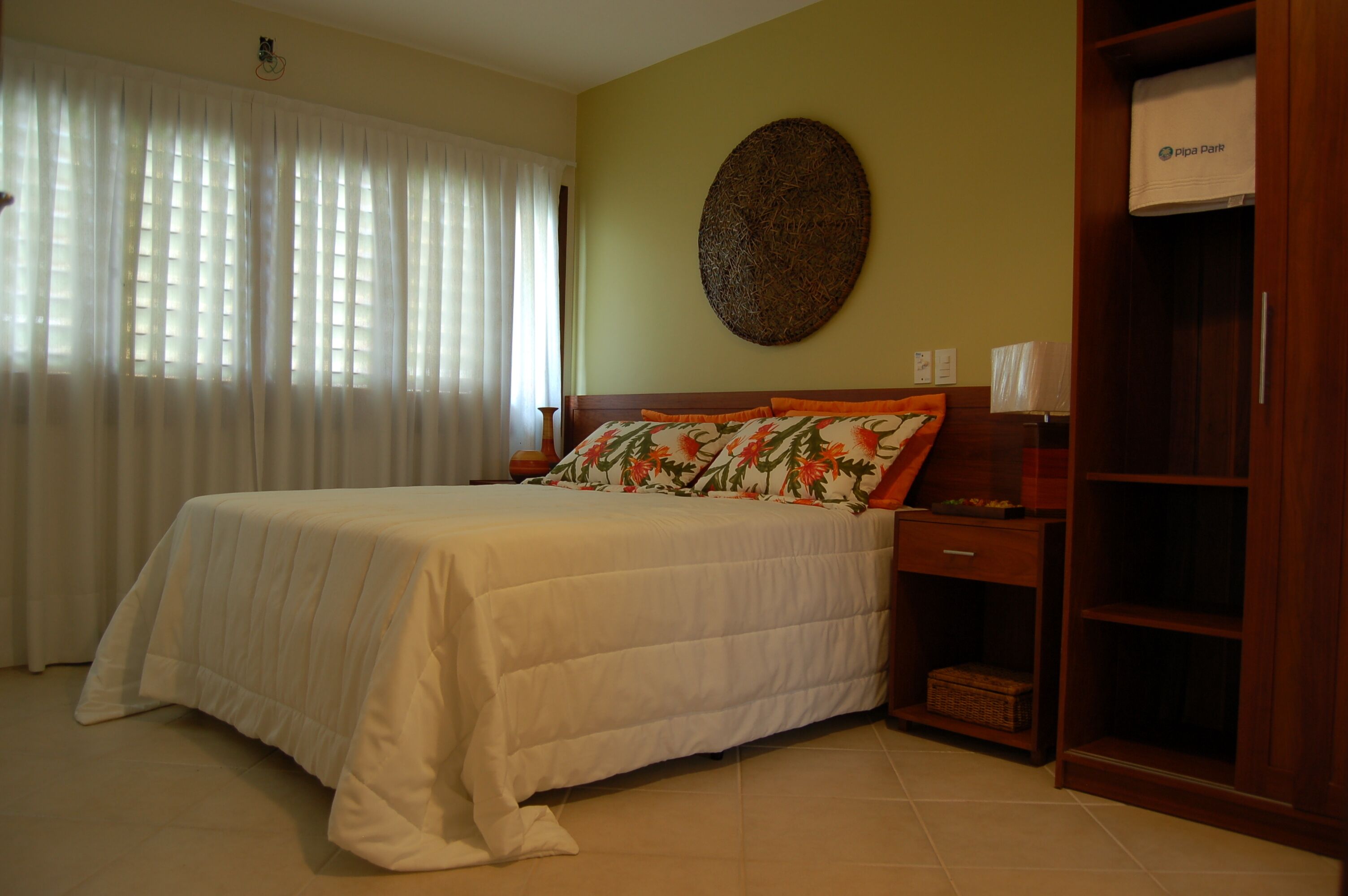 A neatly made bed with a floral pillow, a wooden nightstand, and a wardrobe with a towel hanging.
