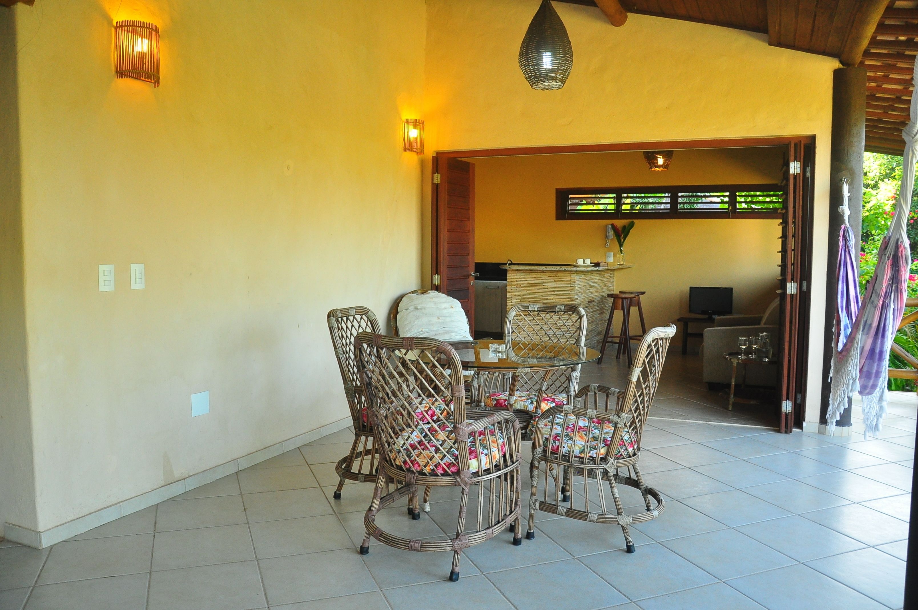 A dining area with wicker chairs and a table, a kitchen in the background, and a covered outdoor area with a hanging light fixture.