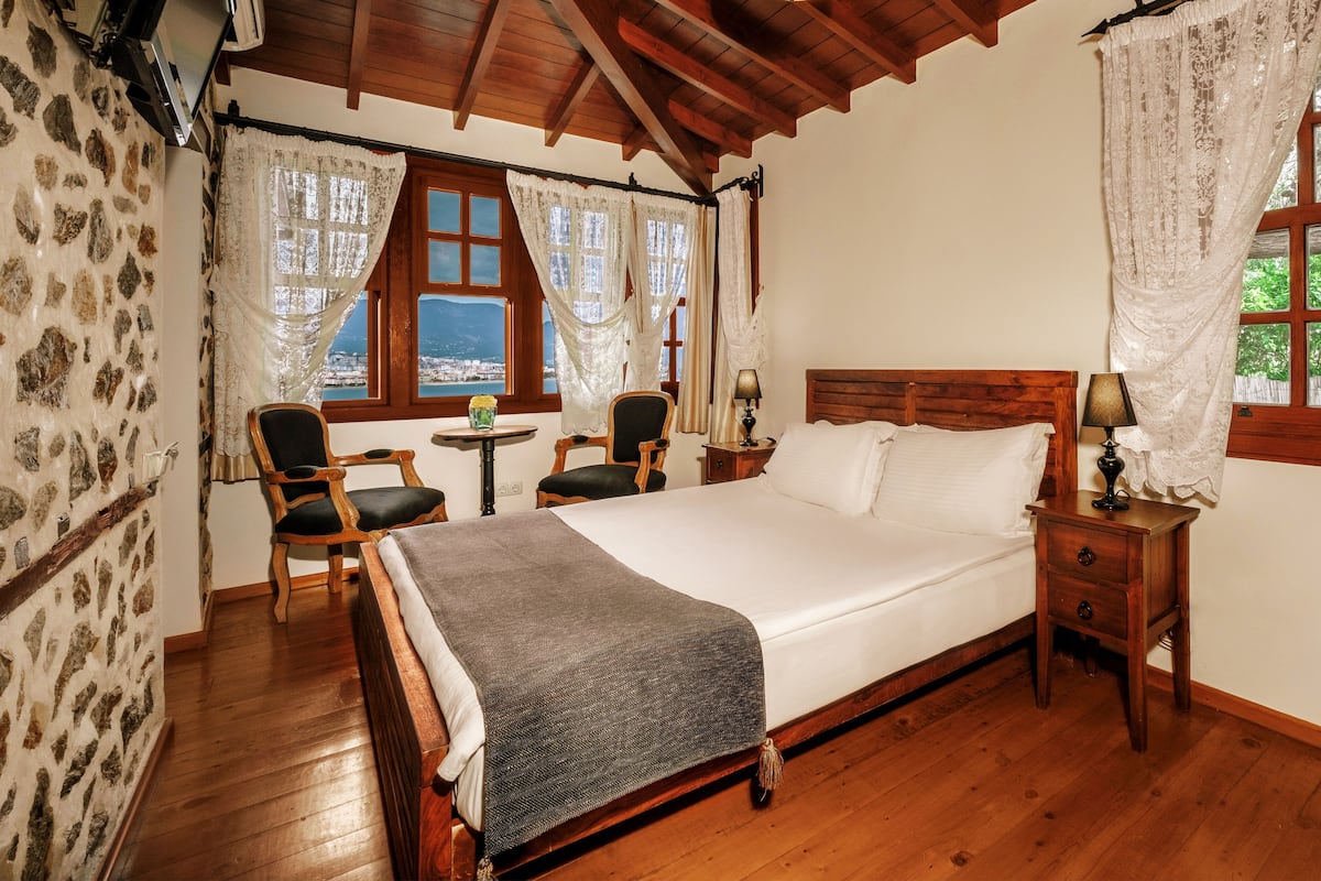 A wooden-framed bed with a white mattress and a gray blanket, a wooden nightstand with a lamp, a wooden chair, and a small table with a vase and a plant.