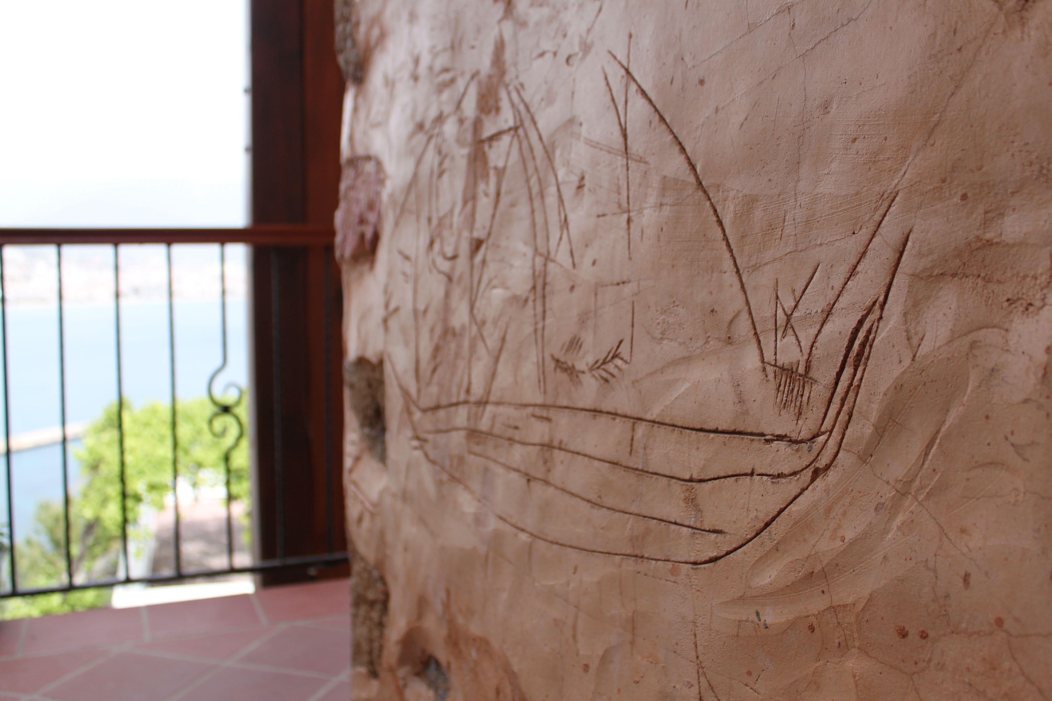 Un gros plan d’un mur à la texture particulière, orné de motifs gravés, un balcon avec une rambarde en métal, et une vue sur la verdure et l’eau en arrière-plan.
