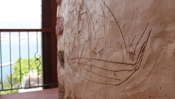Un gros plan d’un mur à la texture particulière, orné de motifs gravés, un balcon avec une rambarde en métal, et une vue sur la verdure et l’eau en arrière-plan.
