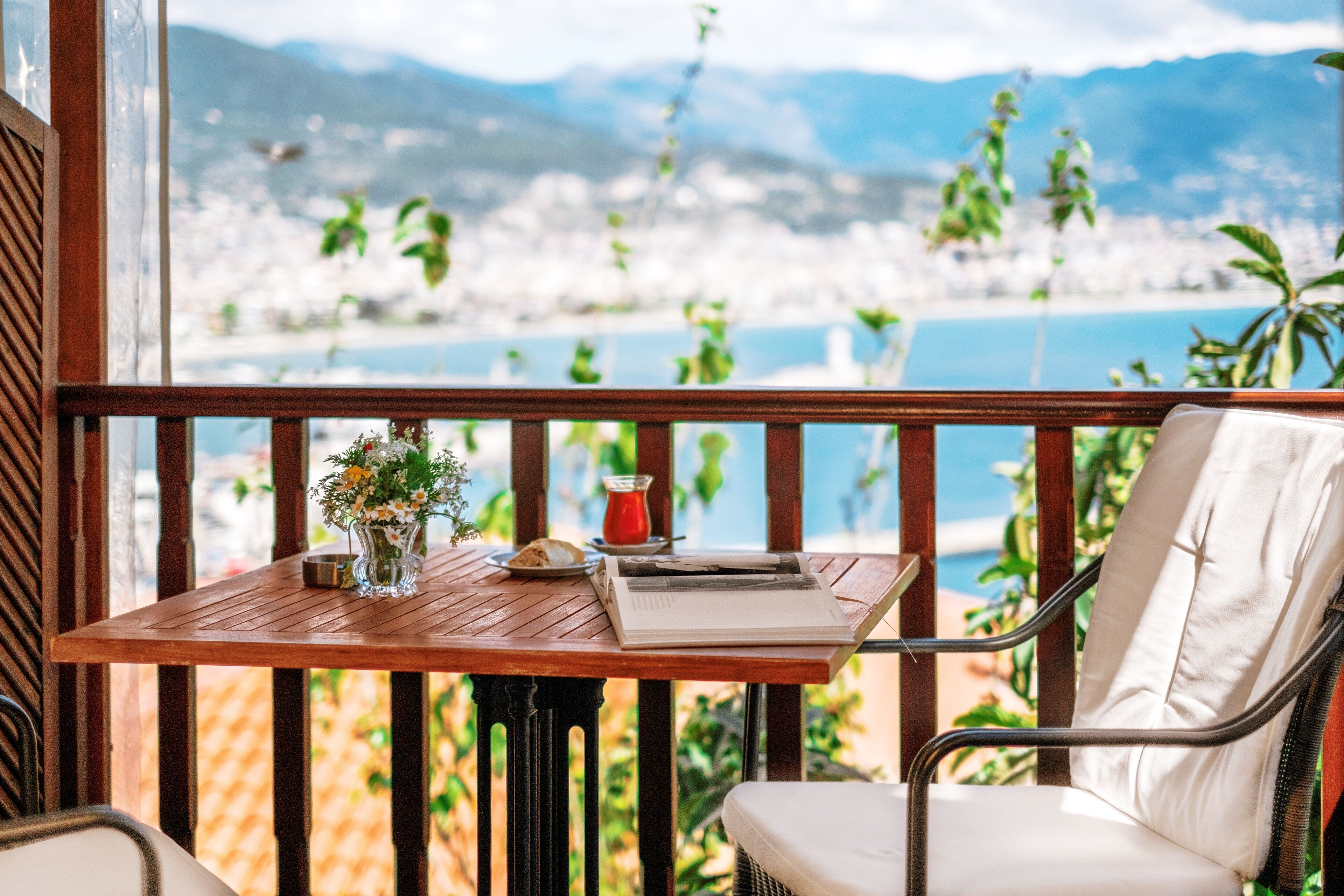 Una mesa de madera con un florero, un libro y un vaso de bebida roja en un balcón con vista al agua y montañas.