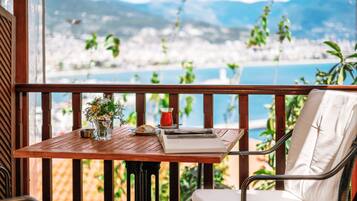 Una mesa de madera con un florero, un libro y un vaso de bebida roja en un balcón con vista al agua y montañas.