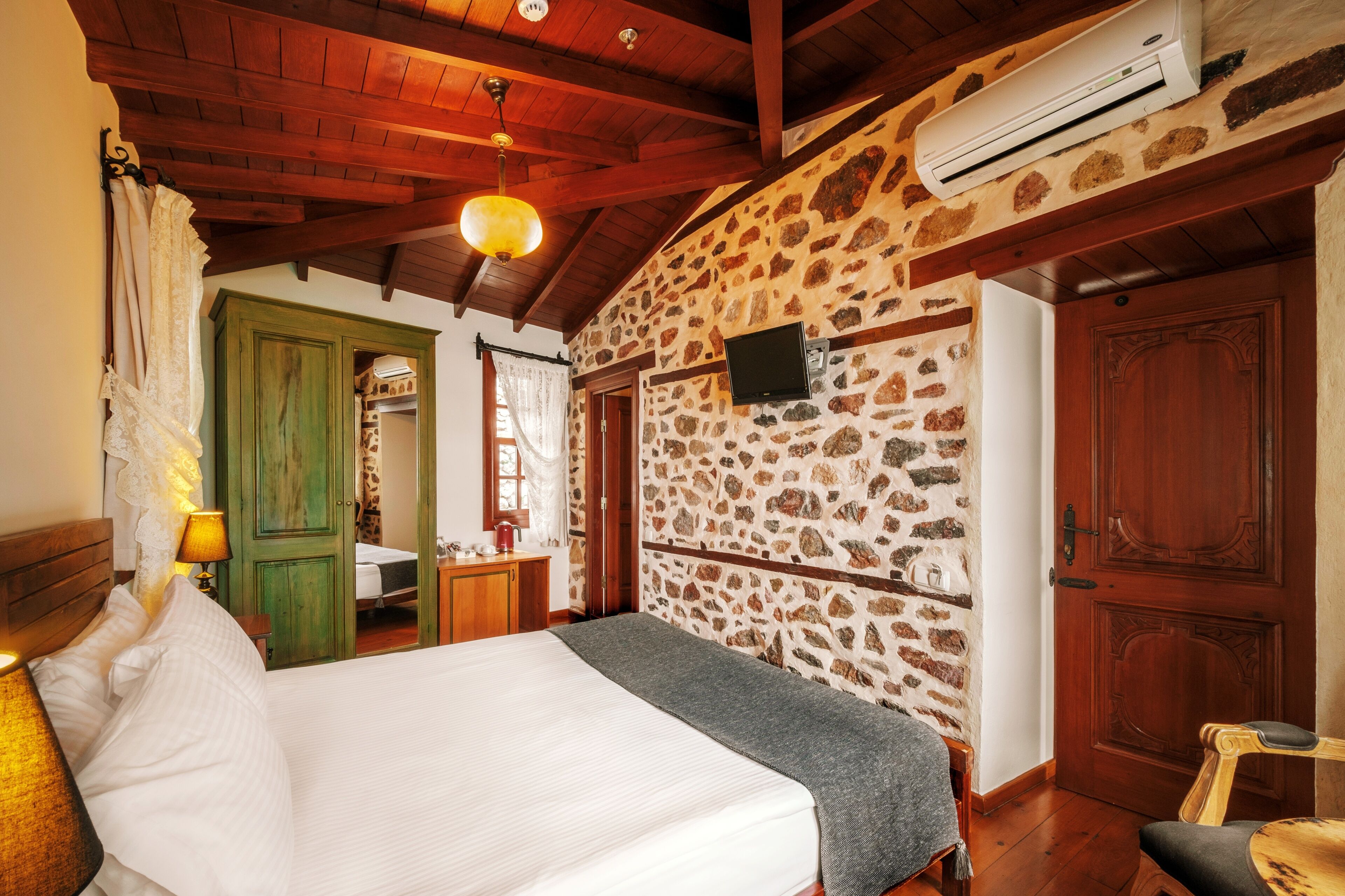 A bedroom with a bed, a wooden ceiling, a stone wall, a TV, and a door.