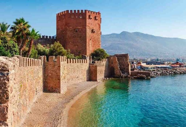 A historic stone tower with a crenellated wall and a clear blue sea in the foreground. - Hotel Villa Turka (Alanya)